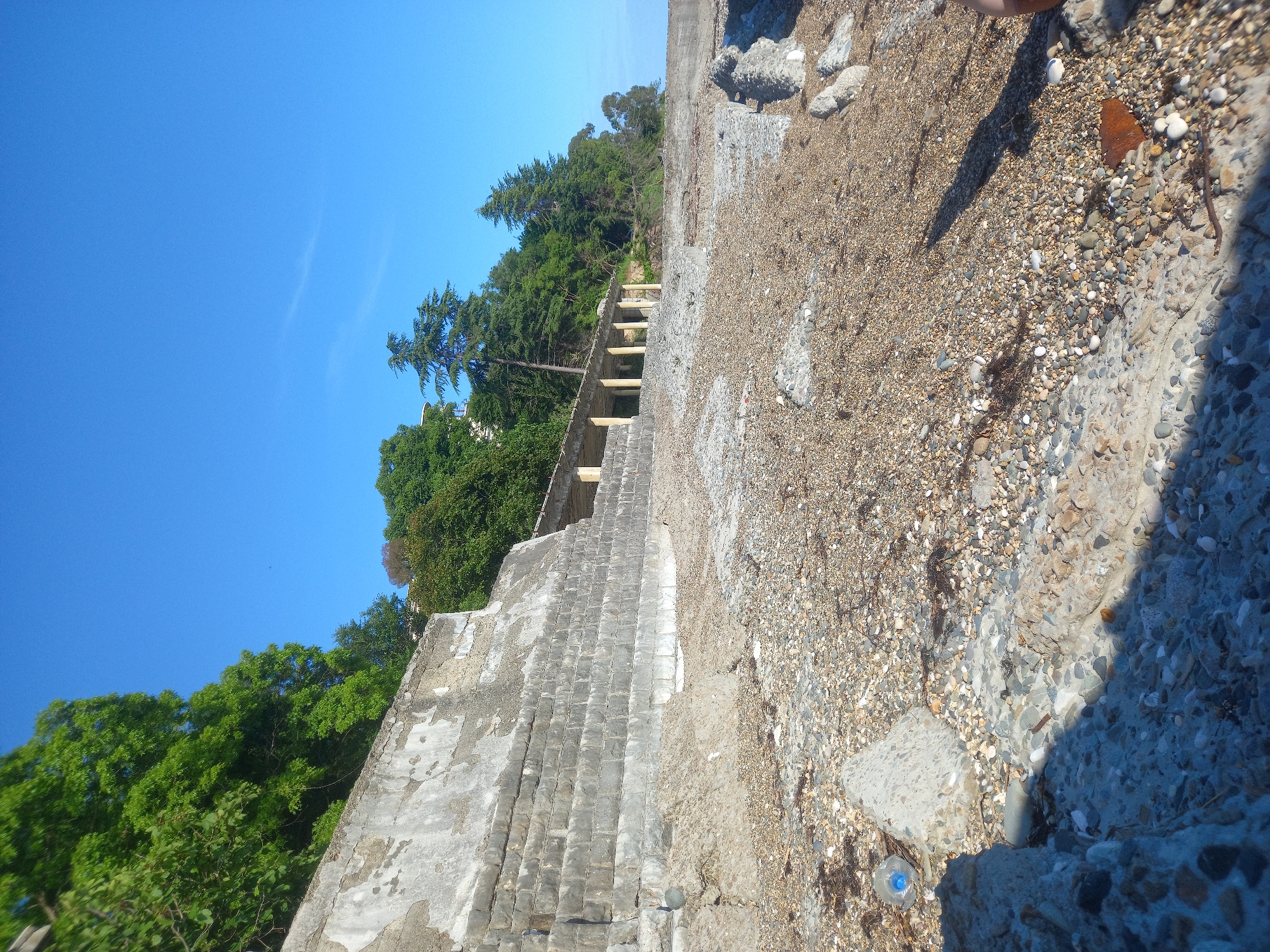 The image depicts a partially constructed or dilapidated concrete structure on a rocky shoreline under a clear blue sky, suggesting a sunny day. The structure appears to be a terraced or stepped embankment made of rough concrete and stone. Green trees and foliage are visible on the upper slopes behind the structure, suggesting a natural, possibly coastal, setting. The ground in the foreground and on the structure itself is covered with small stones and gravel. There's a plastic bottle with a blue cap visible on the ground near the lower left of the image, which is a common litter item. The lighting and shadows indicate it is likely midday. The scene evokes a sense of abandonment or a work-in-progress, with no people visible. The context provided, Gwandra, Abkhazia, places this scene in a specific geographical region known for its coastline and historical sites.