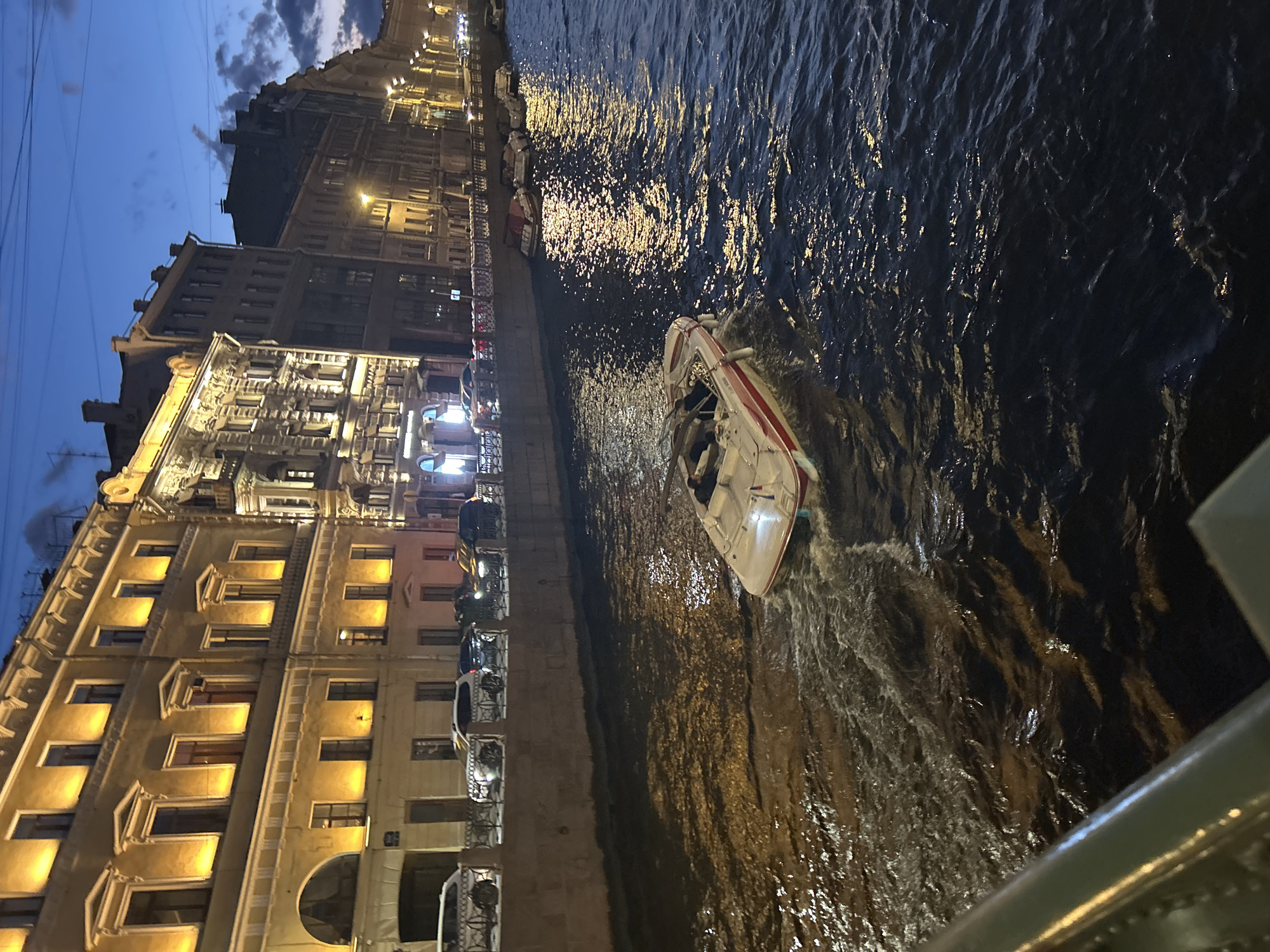 An outdoor, eye-level, medium shot shows a wide boat moving down a dark canal at dusk in an unknown city. The boat is white with a red stripe and has a dark interior. Two people are seated inside the boat, with the person in the driver's seat steering. The water is dark and choppy, with reflections of lights from the buildings on the right. The buildings are tall and ornate, with lit windows. Cars are parked along the canal's edge. The sky is a deep blue with scattered clouds.