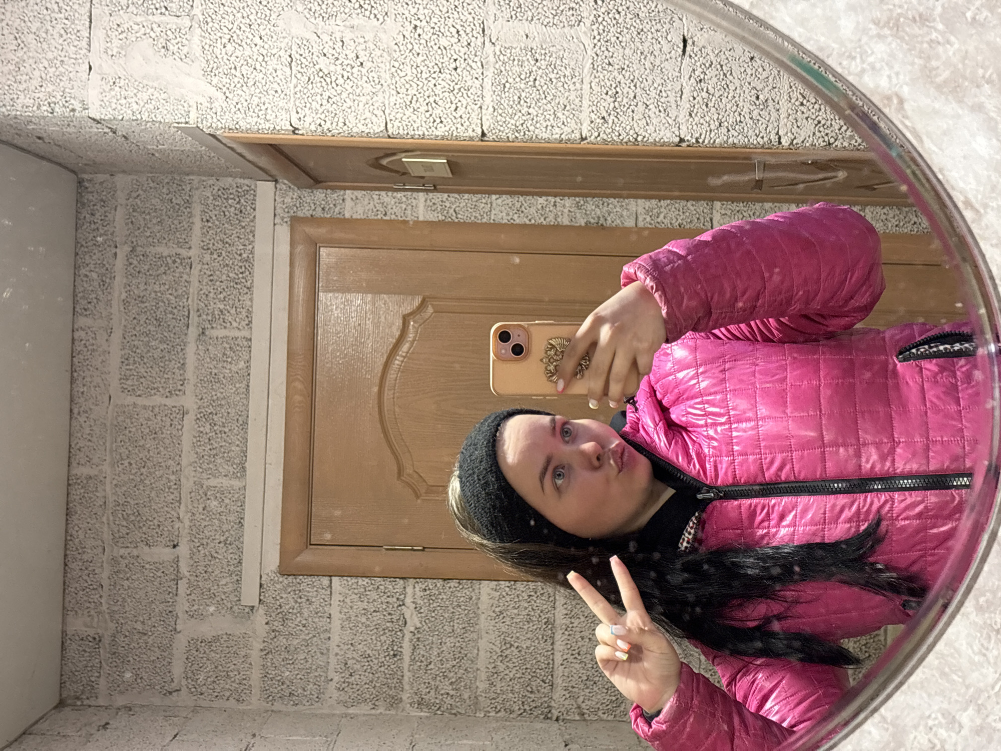 A young woman is taking a selfie in a mirror. She is wearing a pink quilted jacket and a black headband, and she is holding a cell phone. She is making a peace sign with her fingers. The background consists of a wall made of cinder blocks and a door. The location is Русский Вожой, Russia.
