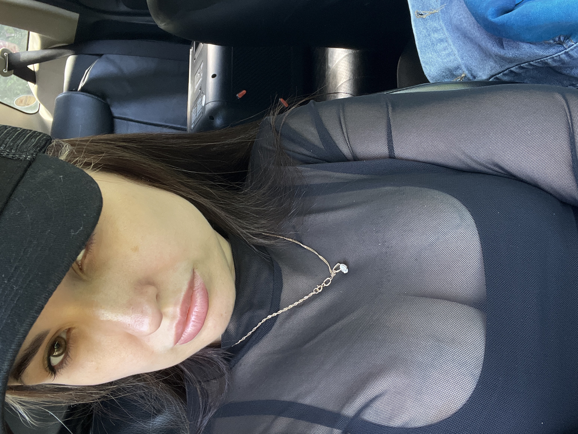 A woman with dark hair wearing a black mesh top and a black cap is sitting in a car. She has fair skin and a necklace with a small pendant. The car's interior includes black leather seats and part of the dashboard. A blue denim jacket is partially visible near the top of the frame. The image appears to be taken from a slightly elevated angle looking down at the woman. The location is unknown.