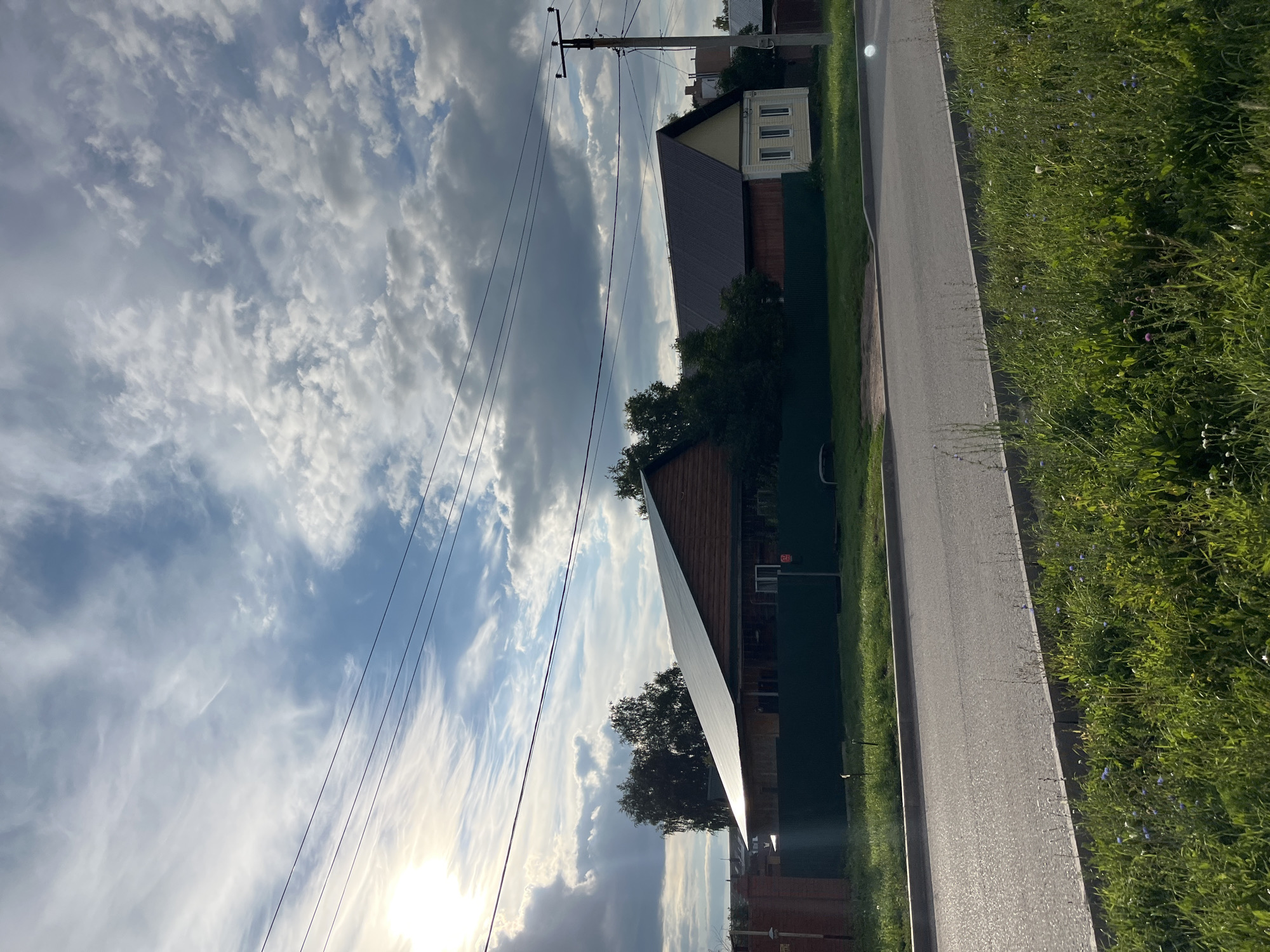 The image shows a scene in Ufa, Russia. A two-story house with a dark roof and wooden siding is visible. A green fence runs along the property line. The house is situated on a street with a paved road and a grassy area with wildflowers. The sky is cloudy with patches of sunlight. Power lines and a utility pole are also present in the scene. The image appears to be taken during the daytime.