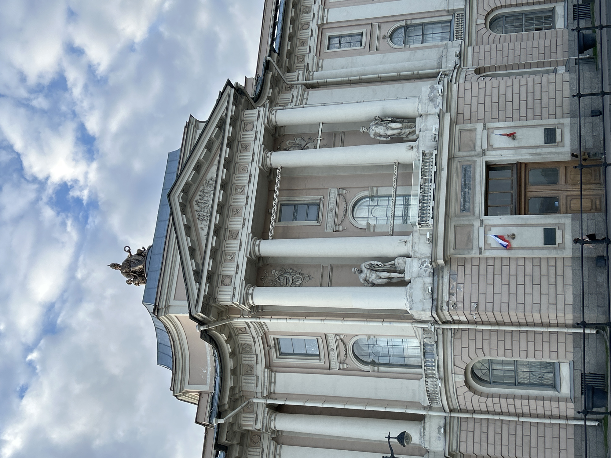 The image depicts the facade of a classical architectural building, likely located in Saint Petersburg, Russia. The building has a pale pink brick facade and is decorated with white columns, sculptures, and ornate details. There is a statue of a soldier holding a shield on the top of the building. The windows are arched, and there is a Russian flag displayed near a doorway. A few dark horizontal poles are visible on the right side of the image. The sky above is cloudy.