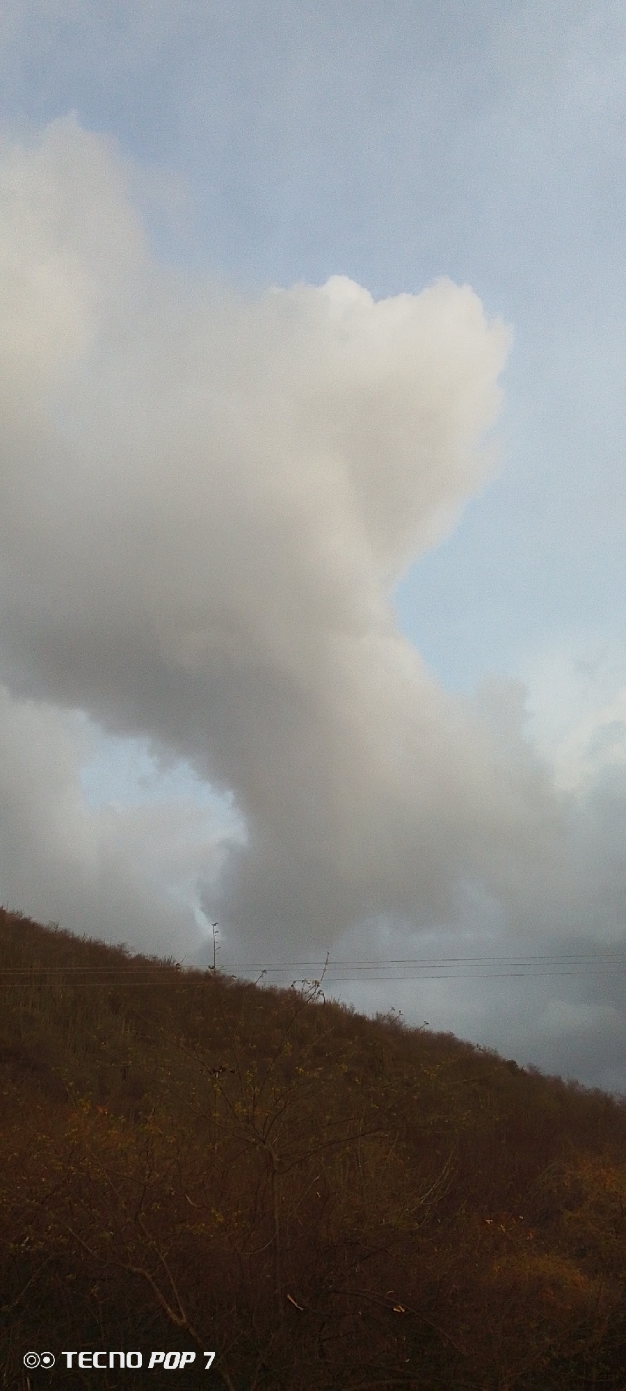The image shows a hillside covered in brown vegetation under a cloudy sky in Altagracia, Venezuela. The sky is mostly obscured by large, gray clouds. A few power lines are visible stretching across the hillside. The vegetation on the hill appears dry. A "TECNO POP 7" watermark is visible in the bottom left corner of the image.