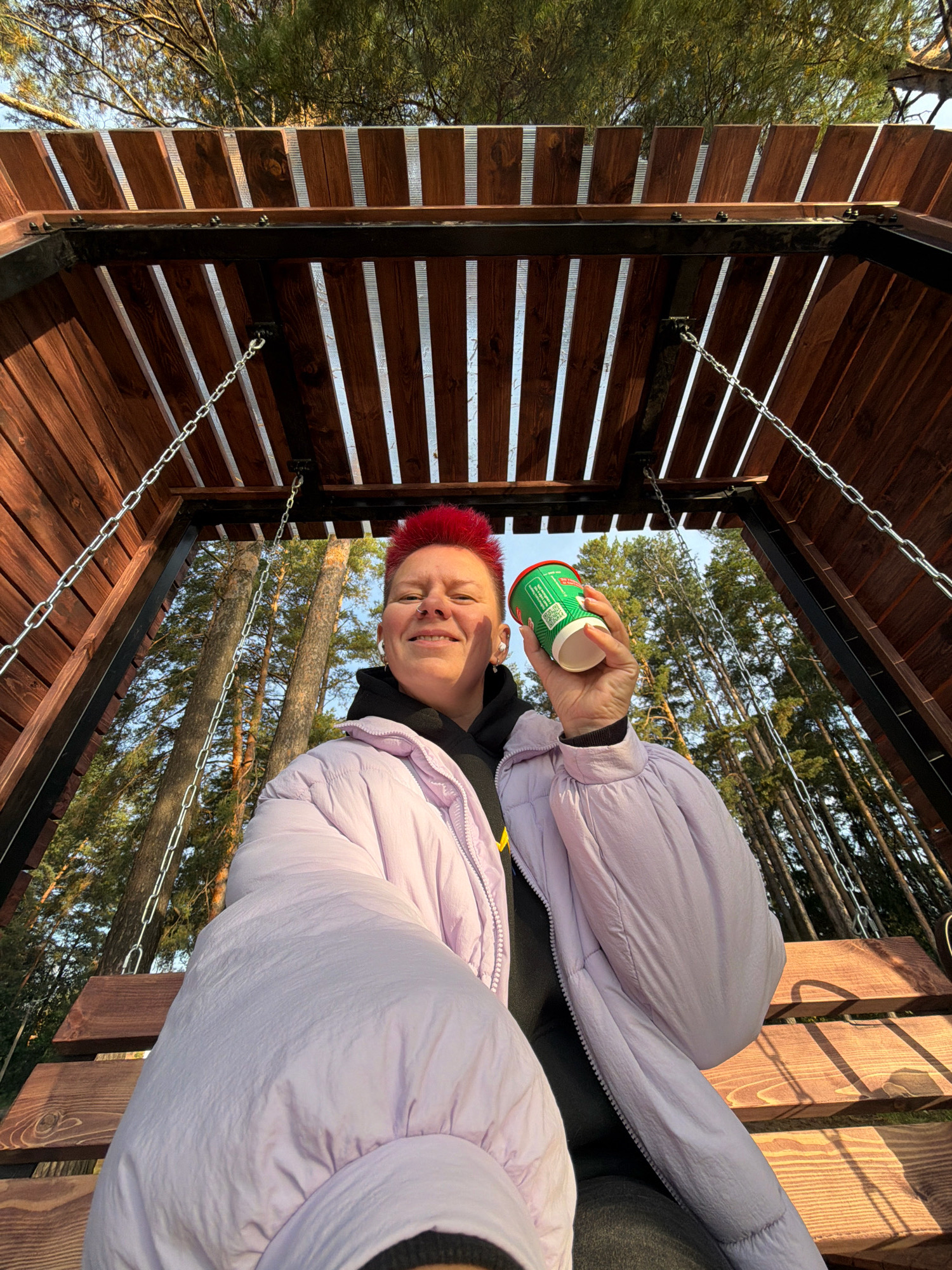 A person with bright red, spiky hair and a nose ring is sitting on a wooden swing seat, holding a green and white paper cup in their right hand. They are wearing a light purple puffer jacket over a black hoodie. The swing is suspended by chains from a wooden structure. The background shows a forest with tall pine trees and a clear blue sky. The lighting suggests it is daytime, possibly late afternoon, given the warm sunlight casting shadows. The location is a public recreational area in the city of Chaikovsky, Russia, likely a park or a nature trail.