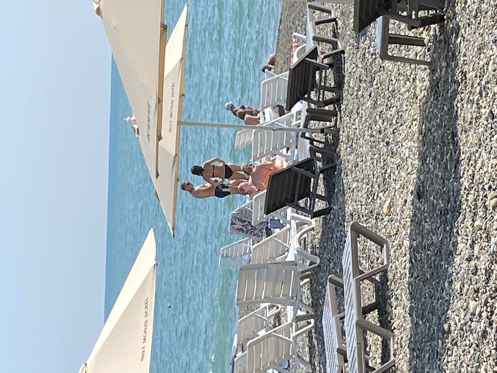 The image depicts a sunny day at a pebbled beach in Chabkwara, Abkhazia. Several people are present, some wading in the turquoise sea, while others recline on white plastic lounge chairs arranged on the shore. Two large cream-colored umbrellas provide shade, one of which has text that appears to read "BRT MORS HOTEL". In the foreground, more lounge chairs are visible, some empty and some occupied by beachgoers. The water is calm with small waves gently lapping the shore. The sky is clear and blue, indicating pleasant weather.