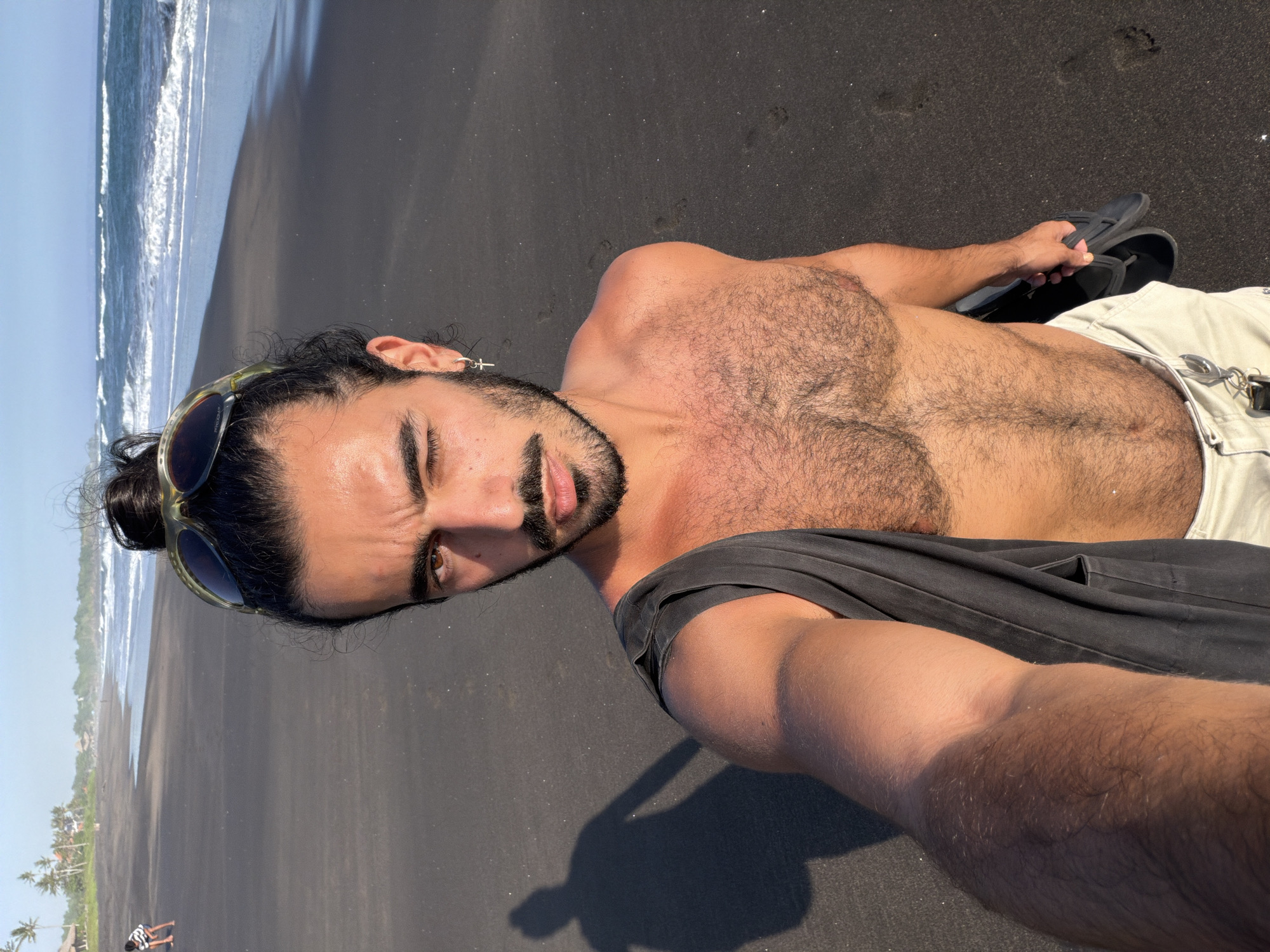 A man with dark, curly hair tied in a bun and a beard is taking a selfie on a black sand beach. He is wearing sunglasses on his head and a dark gray shirt draped over his shoulder. He is also wearing light-colored shorts and holding a pair of black flip-flops. The ocean with waves is visible in the background, and the sky is clear and blue. The man is partially facing the camera, with one eye winked. The shadow of the man is visible on the sand to his left. There are a few people in the distance on the beach. The location is not identifiable as Kediri, Indonesia based on the visual information provided in the image.