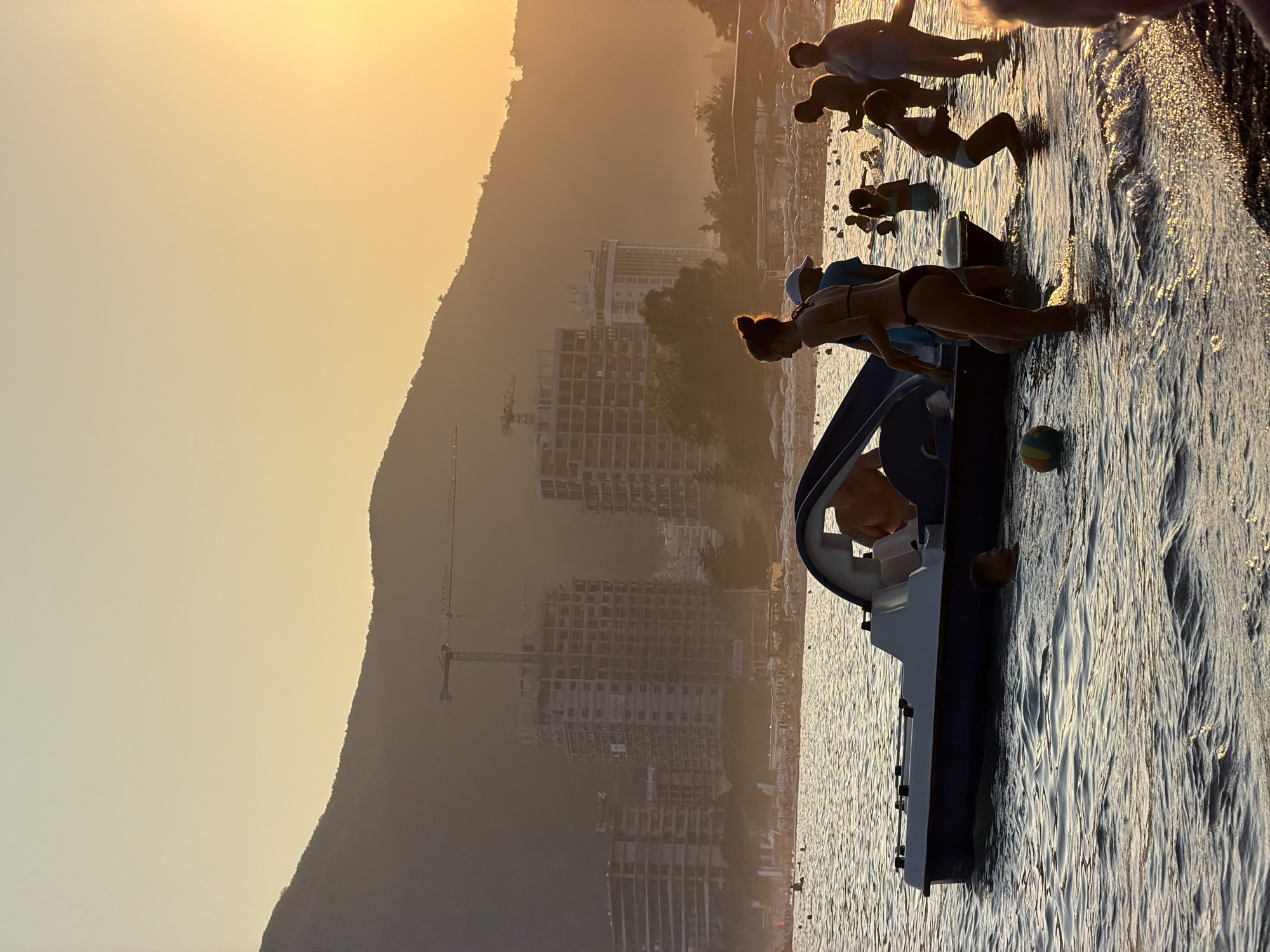 The image depicts a beach scene in Bečići, Montenegro, during sunset. The sky is a warm gradient of yellow and orange, with the sun low on the horizon. In the foreground, several people are visible in the water. A pedal boat is partially submerged, with a person inside and others wading around it. Some individuals are swimming, while others stand near the shore.
In the background, construction is underway on multiple large buildings, some of which are mid-rise and others appear to be high-rise. Construction cranes are prominent above these structures. Beyond the buildings, a hazy mountain range forms the backdrop, with the warm light of the setting sun casting a glow over the entire landscape. Beach umbrellas and awnings are visible along the shoreline, indicating a developed beach area. The overall atmosphere is that of a busy beach at dusk.