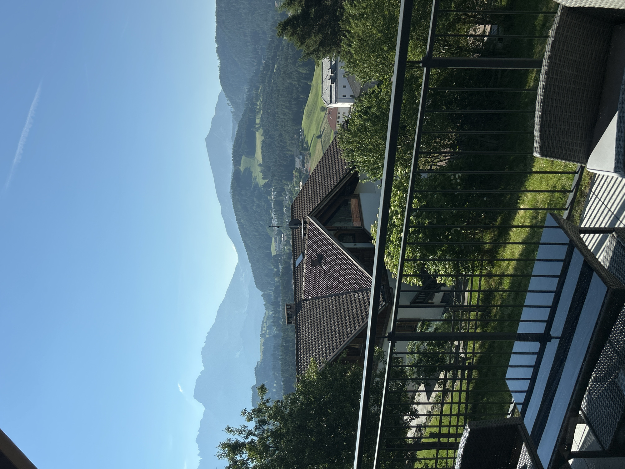 The image displays a panoramic view of a mountainous landscape from a balcony. In the foreground, a balcony railing made of dark metal separates the viewer from the scenery. A table with a dark, possibly glass, top sits in front of a wicker chair. Beyond the balcony, a house with a brown tiled roof is visible, nestled amongst trees. The landscape then unfolds into rolling hills covered in dense green forest, with some lighter green fields interspersed. In the distance, several layers of mountains are visible, fading into a hazy blue in the far background under a clear, bright blue sky. A faint white contrail is visible in the sky. The overall scene suggests a peaceful, rural or mountainous setting, likely during the daytime with good weather.