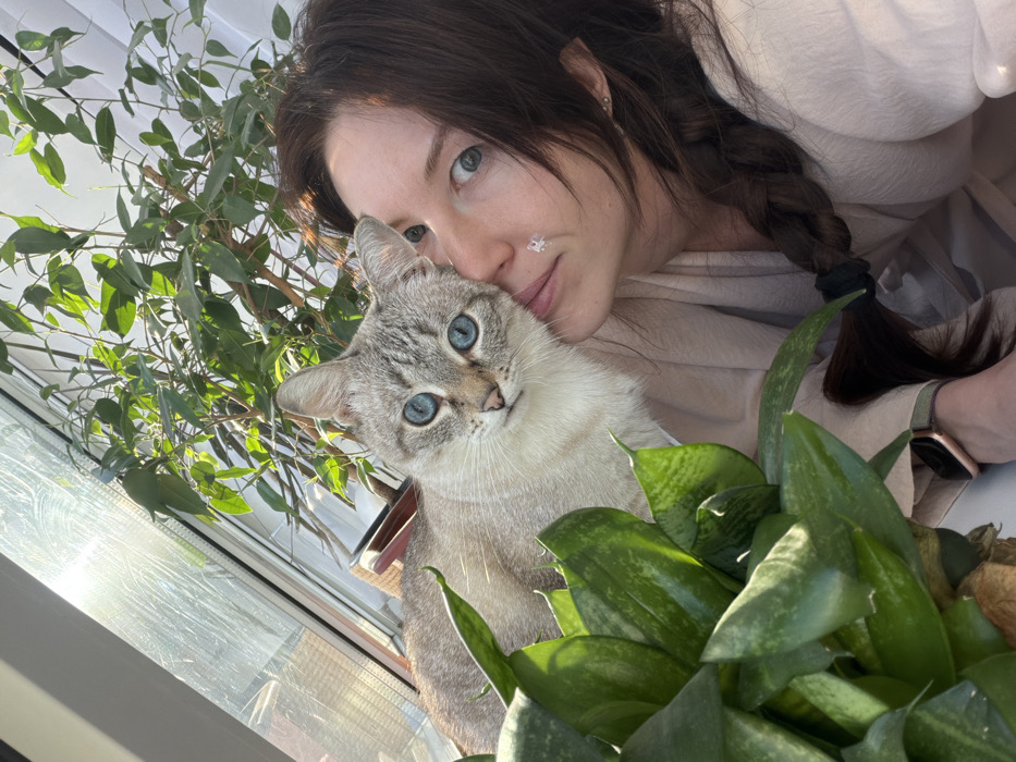 A light-skinned woman with a dark braid leans in next to a light gray cat with striking blue eyes. The woman has a small sticker on her cheek. They are positioned in front of two plants. The left plant has small, round leaves, while the right plant has long, pointed leaves. Sunlight shines through a nearby window. The woman is wearing a loose, light-colored garment, and a watch is visible on her left wrist. The image is taken from a slightly elevated angle, capturing a sense of warmth and intimacy.