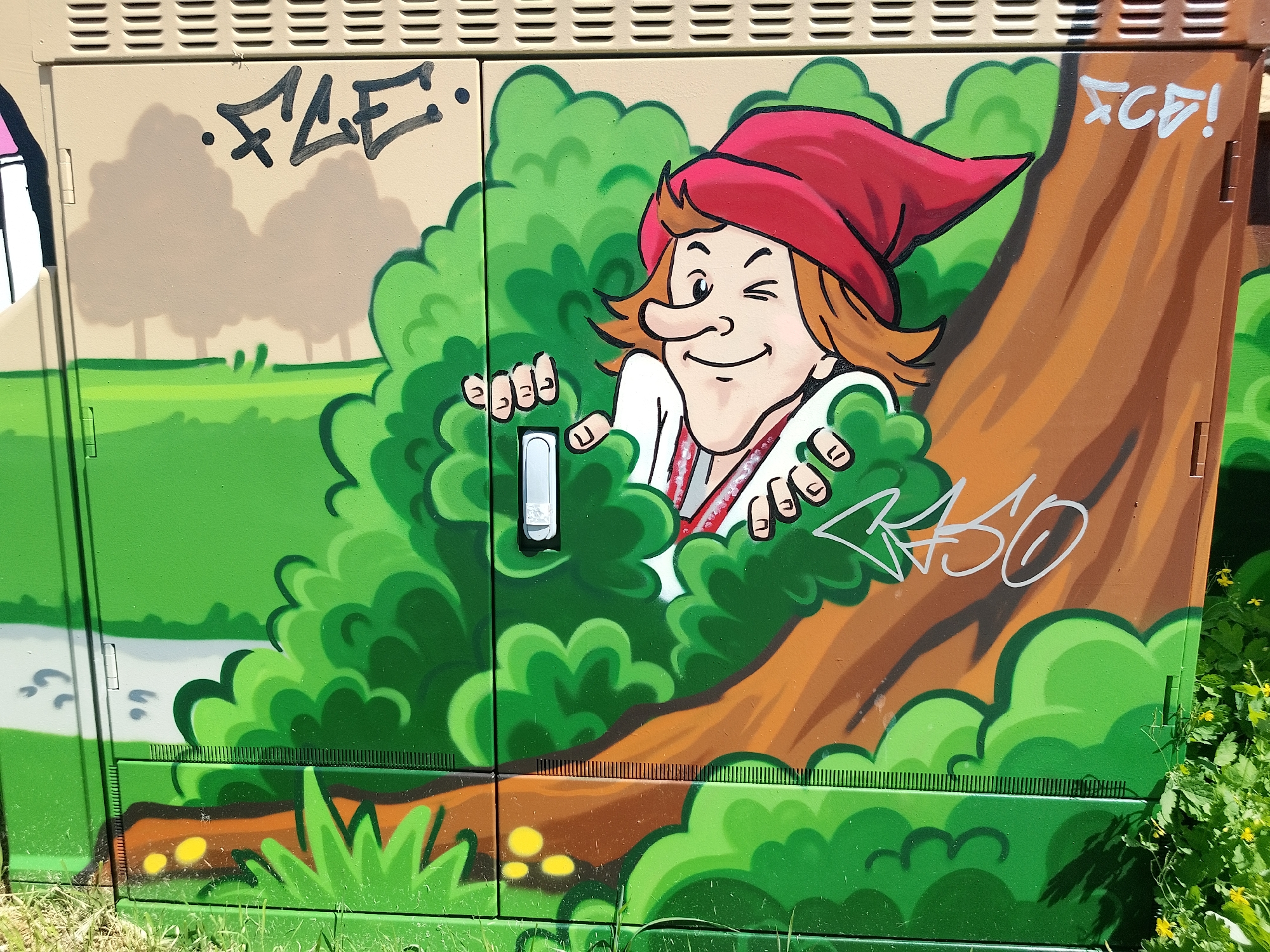 A medium-shot photograph captures a whimsical cartoon mural painted on a utility box in Unknown, Unknown. The artwork features a fair-skinned cartoon figure with a winking expression, wearing a red cap and traditional white shirt with red details, partially obscured by lush green foliage. The figure's hands are playfully positioned at the edges of the leaves, as if peeking out from behind them. A brown tree trunk provides a backdrop to the right of the figure, and a lighter brown landscape with green grass is visible in the background on the left. Graffiti tags in black and white are scrawled across various sections of the box. The lower portion of the box depicts more green grass and foliage, blending with the real grass and weeds growing at the base of the utility box. The scene is brightly lit, suggesting it was taken during daylight hours.