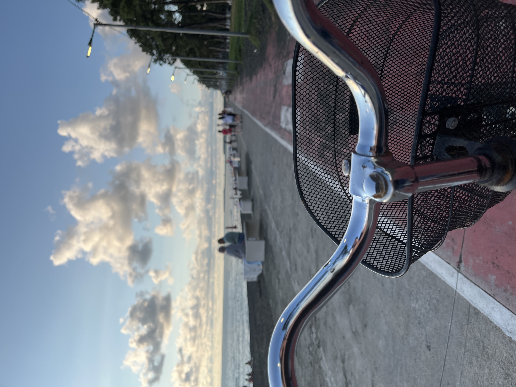 The image captures a perspective from a bicycle on a paved path next to the sea in Ureki, Georgia. The path is marked with red and white lines, suggesting a designated cycling or pedestrian area. On the left, the sea is visible, with gentle waves lapping the dark sand beach. Several people are seated on benches along the beachfront. In the distance, more individuals are walking along the path, some dressed in bright clothing. To the right of the path, a row of trees lines the walkway, and streetlights are spaced intermittently. The sky above is a pale blue, adorned with scattered, fluffy white clouds, indicating it is likely late afternoon or early evening. The foreground is dominated by the handlebars and front basket of the bicycle, rendered in chrome and black mesh, respectively.