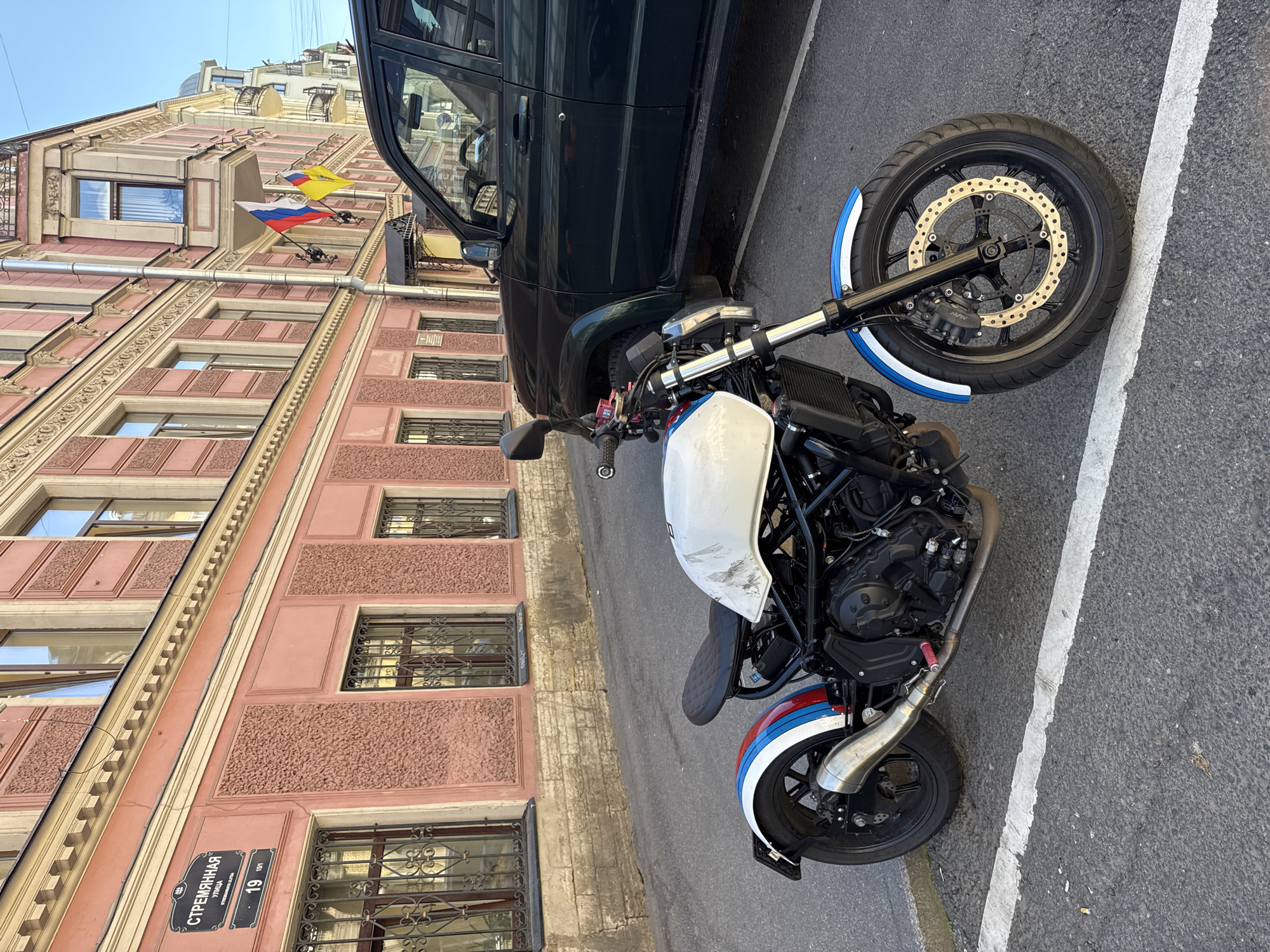 This media file shows a street scene in Saint Petersburg, Russia, on a bright, clear day. The foreground features a motorcycle parked next to a dark-colored SUV. The motorcycle is white with red, white, and blue racing stripes on its front fender. The SUV is dark and partially obscures the building behind it.
The background is dominated by a tall, pink building with ornate architectural details. Multiple windows with decorative wrought-iron grilles are visible. On the side of the building, three flags are displayed: the Russian flag (white, blue, red horizontally), and two other flags, one yellow and one possibly blue and white. A street sign in Cyrillic text, reading "СТРЕMЯННАЯ", "УЛИЦА", and "19", is also visible on the building.
The scene appears to be during daylight hours, suggested by the strong shadows and bright sunlight. The overall mood is neutral, simply documenting the objects and setting.
