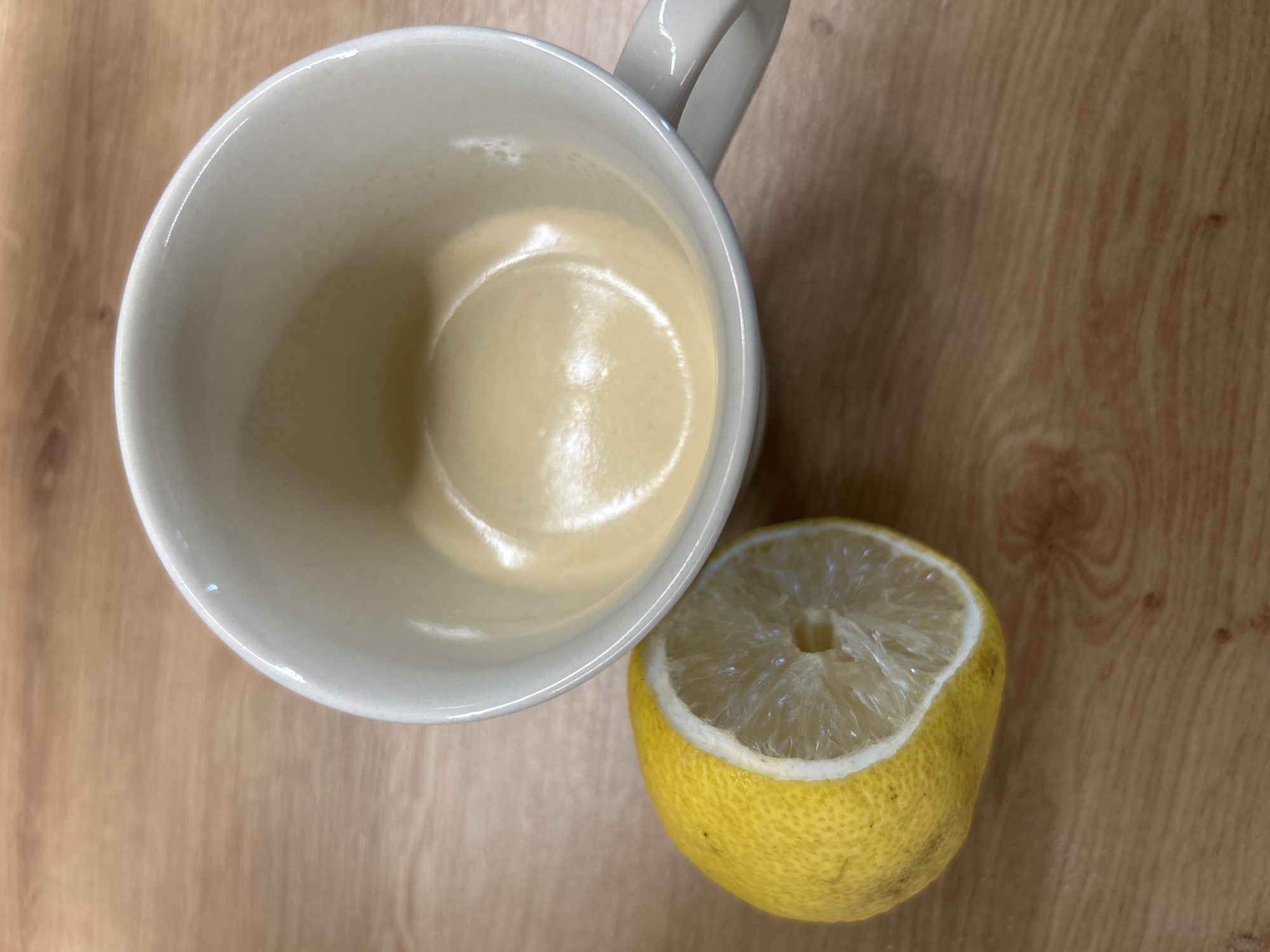 An overhead shot presents a still life composition of a white ceramic mug and a half-cut lemon on a light brown wooden surface. The mug, occupying the upper left and central portion of the frame, is seen from above, revealing a small amount of light, creamy yellow liquid at its bottom, possibly coffee with milk or tea. Reflections from an unseen light source are visible on the liquid's surface and the mug's rim. A portion of the mug's handle is visible on its right side.
To the right and slightly below the mug lies a yellow lemon, cut horizontally. Its exposed, juicy pulp and segments face upwards, while the textured yellow rind shows some minor natural blemishes. The wooden surface, serving as the background, features distinct grain patterns running predominantly horizontally. The scene appears to be indoors, under ambient lighting that creates subtle highlights on the ceramic and liquid. There is no discernible activity, people, or visible text in the image. The time of day and specific weather conditions are not indicated.