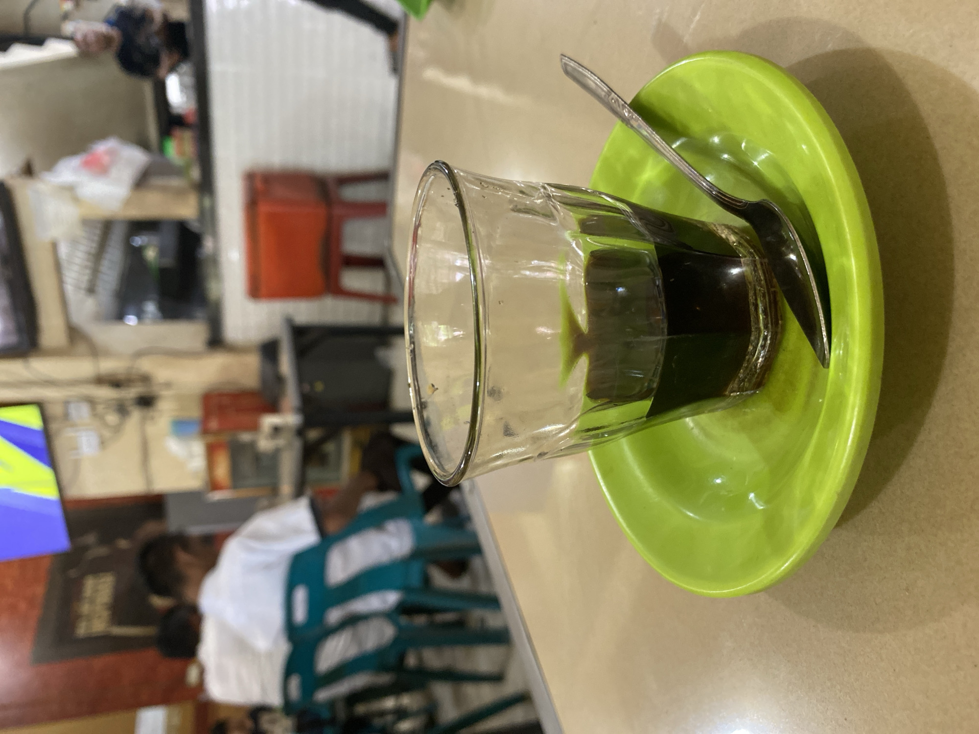 The image shows a close-up of a glass of dark liquid, likely coffee, placed on a bright green saucer. A spoon rests on the saucer, positioned next to the glass. The glass is clear and has some small bubbles visible on its interior surface. The liquid inside is dark and opaque, filling about half the glass. The saucer is a vibrant lime green and has a slightly curved rim. The background is blurred but suggests an indoor setting, possibly a cafe or restaurant.
In the background, there are hints of people and furniture. To the left and slightly out of focus, there are glimpses of what appear to be people seated or moving, with some wearing white shirts. There are also some shelves with items on them, and red chairs visible in the middle ground. The overall lighting suggests it is daytime, possibly late morning or afternoon, as there is natural light contributing to the illumination.
The setting appears to be casual and somewhat busy, with the blurred background indicating activity. The focus on the drink and saucer suggests the image is about the beverage or a moment of quiet enjoyment amidst a lively environment. There is no visible text in the image. The location context is Lhokseumawe, Indonesia, which might imply a local coffee culture or establishment.