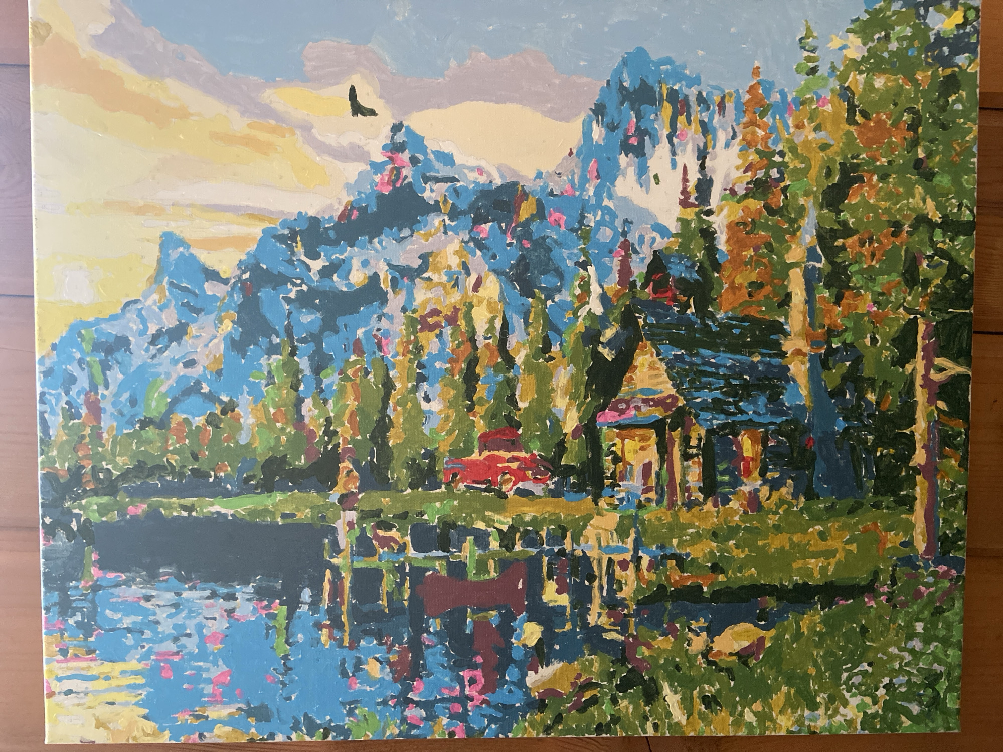 The image is a painting depicting a mountain landscape with a body of water in the foreground. In the background, jagged, blue mountains dominate the scene. A cabin is nestled among the trees. The water reflects the mountains, trees, and the cabin, creating a mirrored effect. A red car is parked near the cabin. The sky is a light blue color with patches of yellow and white. There is a silhouette of a bird flying in the sky. The location is Unknown, Unknown.