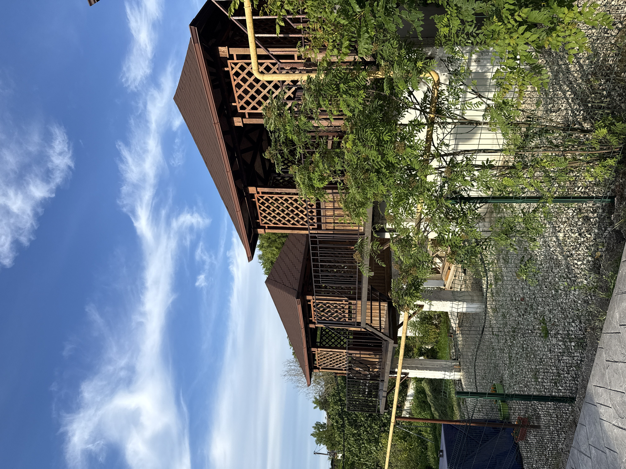 This image, presented as if correctly oriented, captures an outdoor scene dominated by two elevated wooden gazebos or structures under a bright blue sky. The structures feature brown tiled roofs, intricate latticework railings, and dark wood beams, suggesting a rustic architectural style. White support pillars are visible beneath them.
Lush green foliage, consisting of various trees and shrubs, surrounds and partially obscures the gazebos, creating a natural and verdant setting. In the background and to the left, a vibrant blue sky stretches across the frame, adorned with scattered, wispy white clouds.
No people are visible, and no specific activity is taking place, giving the scene a tranquil and serene atmosphere. A yellow pipe runs along one of the structures, disappearing into the greenery. In the foreground and to the right, a paved area with a textured, possibly stone, surface is visible, alongside a chain-link fence. Beyond the fence, a ground cover of gravel or crushed stone can be seen.
The scene depicts a sunny day with clear weather, likely in a recreational or residential area. There is no visible text in the image. The context provided indicates the location is Taman, Russia.