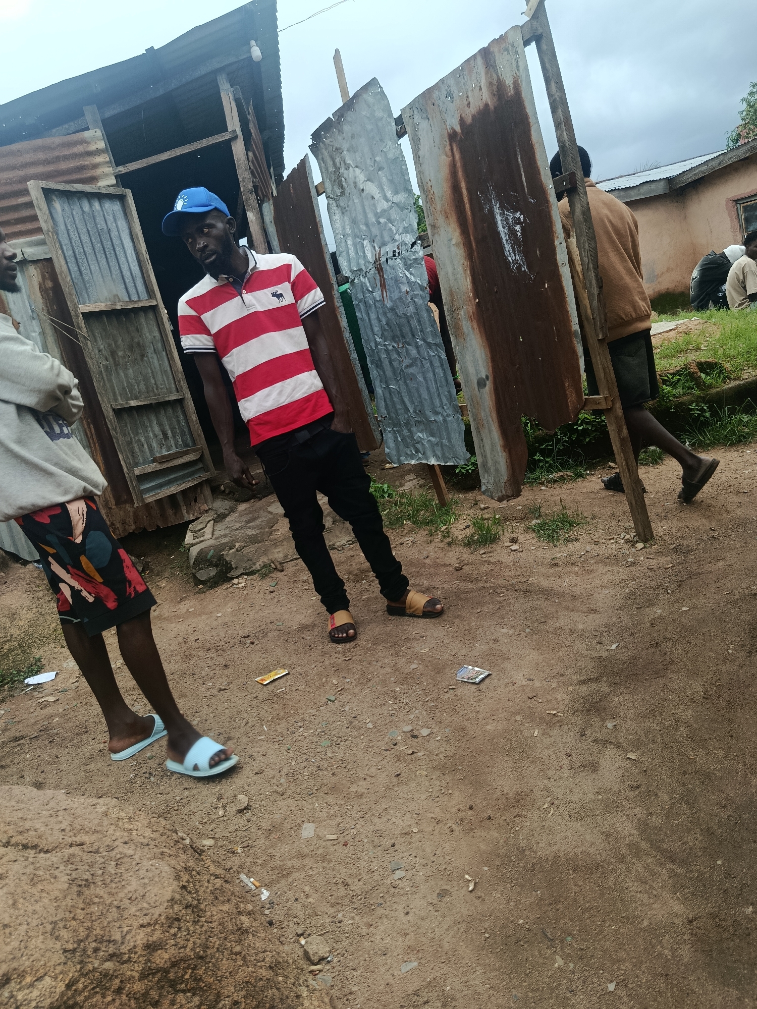 In this medium shot, taken outdoors in what appears to be a rural or underdeveloped urban area, three individuals are visible. The foreground is dominated by a dirt and gravel ground surface, with a large rock formation on the left.
A man, centrally positioned, is wearing a red and white striped polo shirt and black pants. He has a blue baseball cap on and is looking slightly off-camera to the left. His right hand is in his pocket, and his left hand is relaxed by his side. He is wearing brown sandals.
To the left of the central figure, another person is seen from the waist down, wearing a grey hoodie and patterned shorts. They are wearing white flip-flops and their body is angled away from the camera.
In the background, a third person is partially visible on the right side of the frame, wearing a brown jacket and dark pants, with their back to the camera. They are walking away. Behind them, a building with a light-colored wall and a corrugated metal roof is visible. The scene is characterized by makeshift structures constructed from corrugated metal and wood. The sky is overcast.