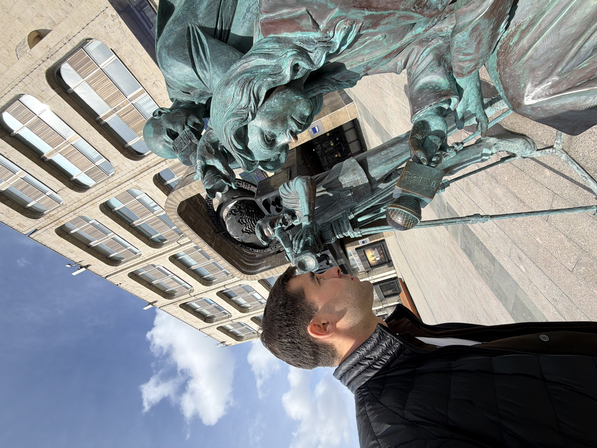 A man in a black jacket looks up at a bronze sculpture. The sculpture depicts three people. One person is in the center holding a microphone labeled "TASS" and looking down. To the left is another person with a bare head. To the right is another person looking down and holding some kind of equipment. Behind the sculpture is a large building with many windows. The sky above has blue sky and white clouds. This appears to be the sculpture "Reporters" in front of the TASS building in Moscow, Russia.