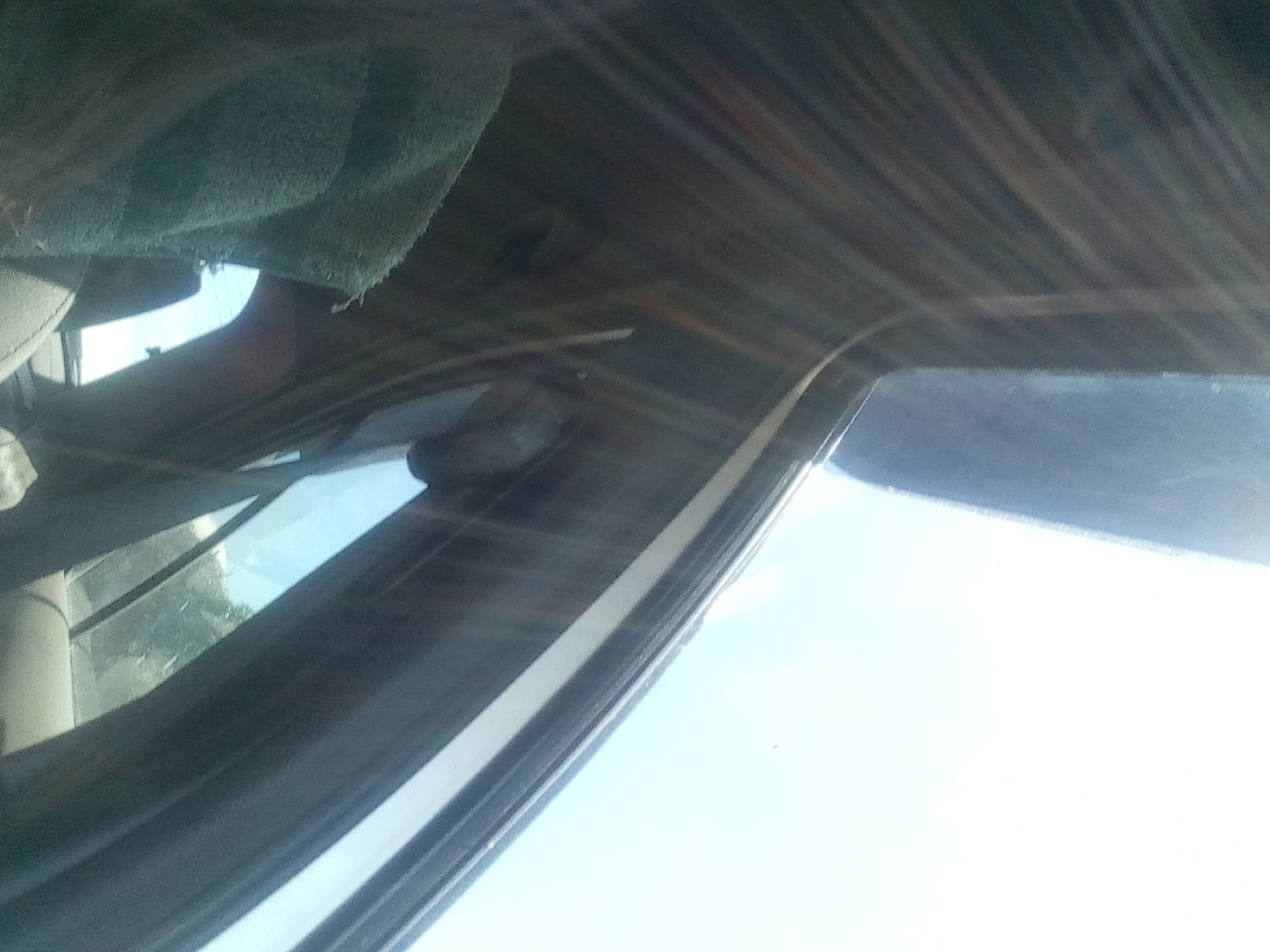 The image appears to be taken from inside a vehicle. The perspective is angled upwards, showing a portion of the car's interior, including what looks like the headliner and a window frame. Sunlight is streaming in, creating strong lens flare effects that obscure details and lend a bright, washed-out quality to the upper portion of the image, which shows a bright sky. A green, textured object, possibly a cloth or blanket, is draped over something in the foreground, partially obscuring the interior. The lighting suggests it is daytime. There is no visible indication of Koviahy, Ukraine, or any urban environment.