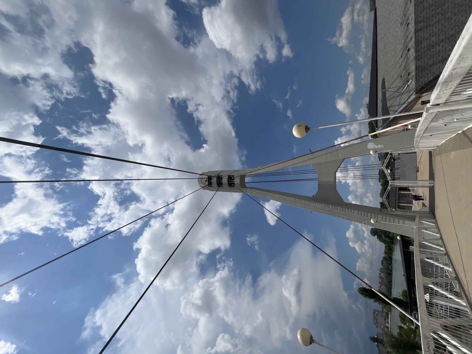 This image captures a view from a modern pedestrian bridge in Krasnodar, Russia, likely the Pedestrian Bridge over the Kuban River. The perspective is looking upwards and forward, emphasizing the bridge's structure and the sky above.
The foreground features the bridge walkway with white railings on either side. The railings are made of metal bars and have some graffiti on them. Two white spherical lamps on poles are visible on the bridge. Further along the walkway, a few individuals are seen walking, including two people in the middle distance.
Dominating the center of the image is a tall, white, A-frame pylon that supports the suspension cables of the bridge. These cables stretch upwards and outwards, disappearing into the vibrant blue sky filled with fluffy white clouds. The pylon has several levels with windows or openings.
Beyond the bridge, a body of water, presumably the Kuban River, is visible, with trees and buildings lining its banks. In the distance, several multi-story apartment buildings are visible. The overall impression is a bright, sunny day with a dramatic sky.