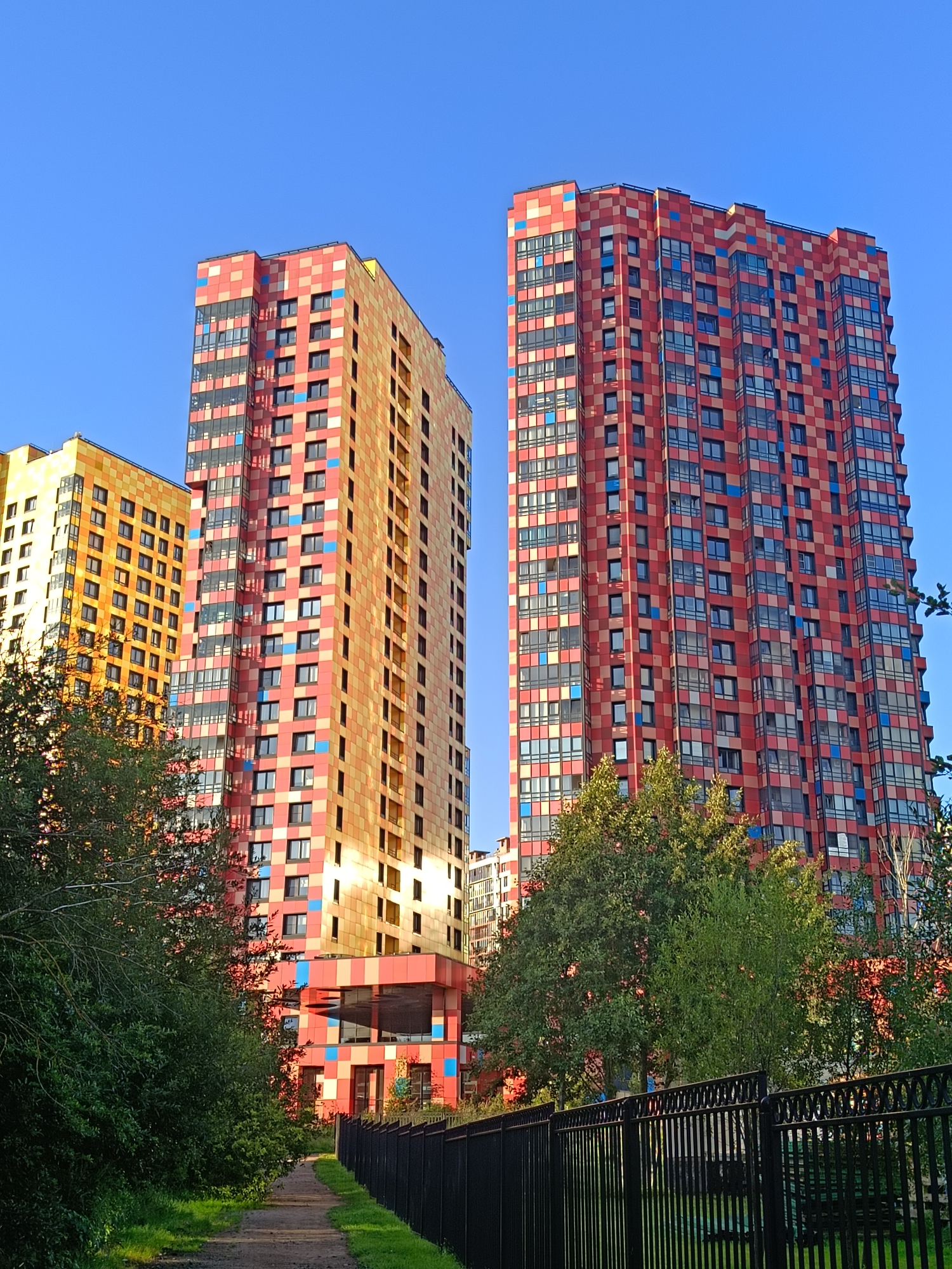 This is a low-angle, eye-level shot of the colorful residential buildings in the "Harmony" residential complex, located in the Primorsky District of Saint Petersburg, Russia. The buildings are characterized by their vibrant facades, featuring a mosaic of red, orange, and blue square tiles, with prominent glass windows.
In the foreground, a dirt path runs alongside a black metal fence, with lush green trees and bushes bordering the path and the fence. The path leads towards the buildings, suggesting an approach to the complex. The sky above is a clear, bright blue. The lighting suggests it is either early morning or late afternoon, with the sun casting a warm glow on the buildings and foliage. The image captures a sense of urban development in a green setting, with the striking architecture of the apartment buildings as the focal point.