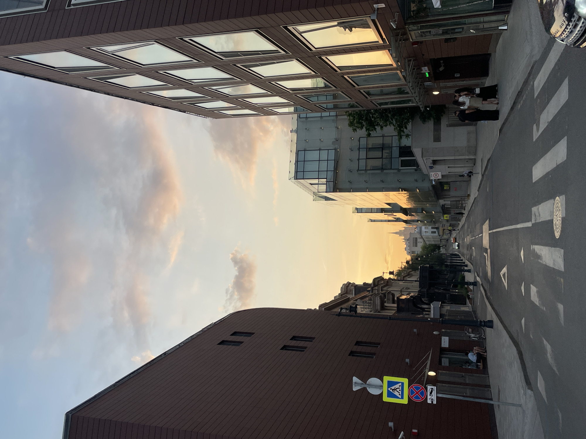 The image depicts a street in Moscow, Russia, framed by buildings on either side. The sky above shows a sunset, with hues of orange and pink. The buildings have a modern architectural style, with one featuring a grid of rectangular windows that reflect the sky. The street below includes a pedestrian crossing, with a group of people gathered on the sidewalk near a building entrance. Street signs and other street furniture are visible, including a sign with a pedestrian crossing symbol and additional traffic signs. The street extends into the distance, with smaller buildings and structures visible under the sunset light.