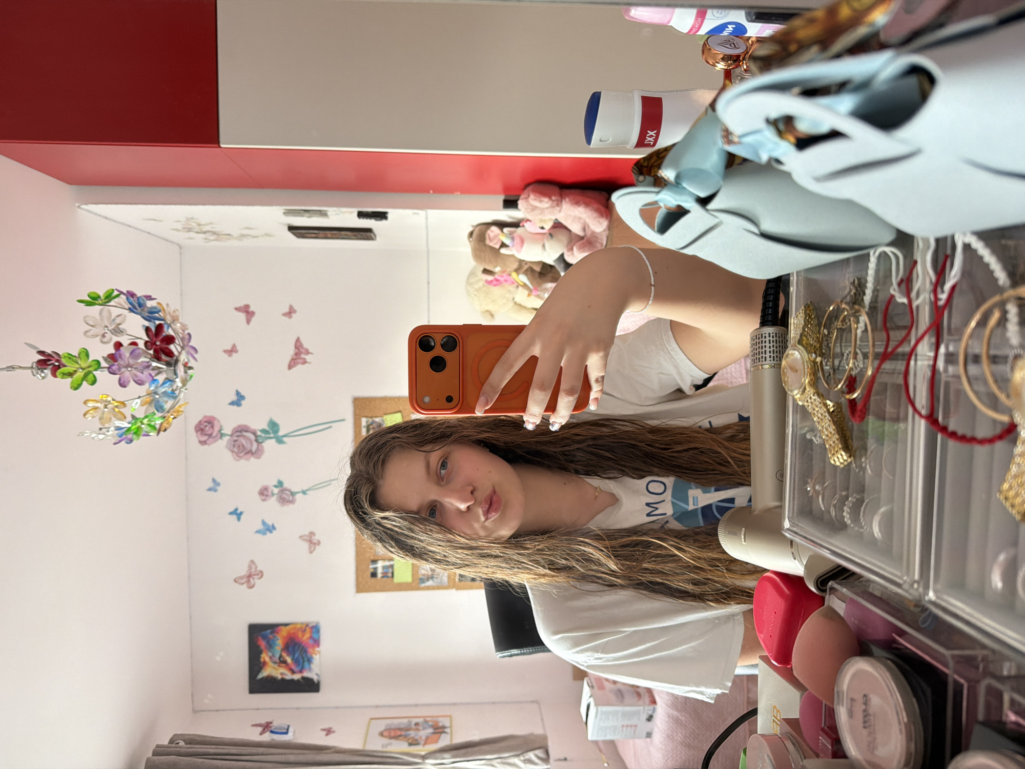 A young woman is taking a selfie in front of a mirror. The mirror reflects a room with flower and butterfly decals on the wall, a colorful hanging decoration, and framed artwork. There is a red and white cabinet above the mirror. The woman is holding an orange smartphone and wearing a white t-shirt. In front of the mirror is a vanity area with cosmetics, jewelry, and a light blue bag. The location is likely indoors, potentially in a residential setting, but there are no explicit indicators confirming Burgas, Bulgaria.