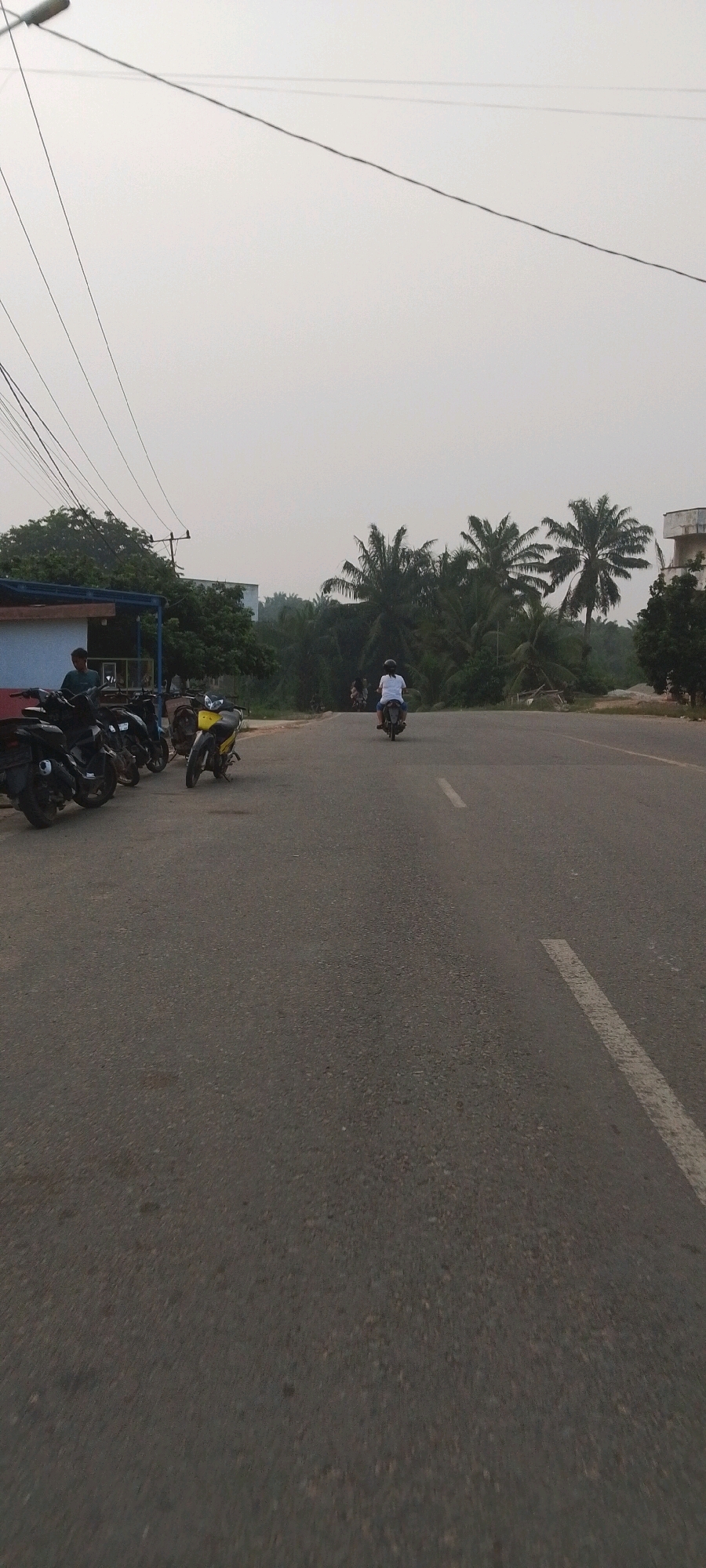 The image shows a street scene with several motorcycles parked on the left side, next to a building with a blue awning. A man in a green shirt stands near the parked motorcycles. Further down the street, a person is riding a white motorcycle away from the camera. In the background, there is dense foliage with palm trees. The sky is overcast. This scene is likely located in Lenggadai Hulu, Indonesia, a region known for its tropical climate and palm trees.
