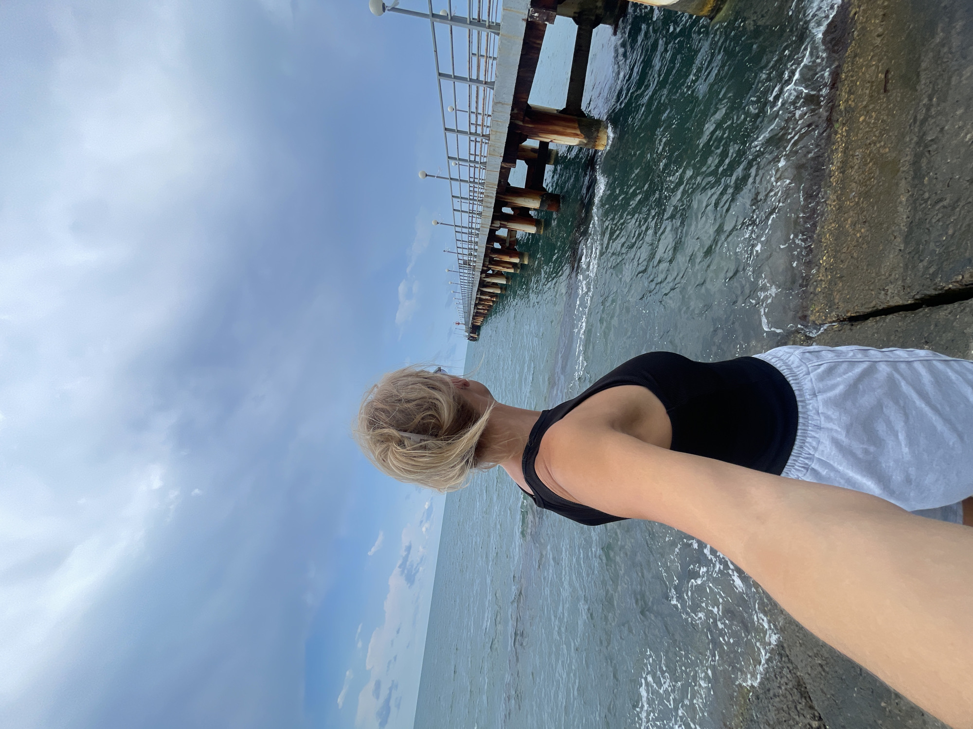 The image shows a woman standing at the edge of a concrete structure, facing the sea. She has blonde hair and is wearing a black tank top and light-colored shorts. The sea is calm with small waves. In the background, a long pier extends into the water, supported by numerous wooden pillars. The pier has a metal railing and is lined with lampposts. The sky is overcast with clouds. The scene appears to be set during the daytime, possibly in the late afternoon or early evening, given the soft light. The location is identified as Sochi, Russia. The woman's posture suggests contemplation or enjoyment of the seaside view. There is no visible text in the image.
