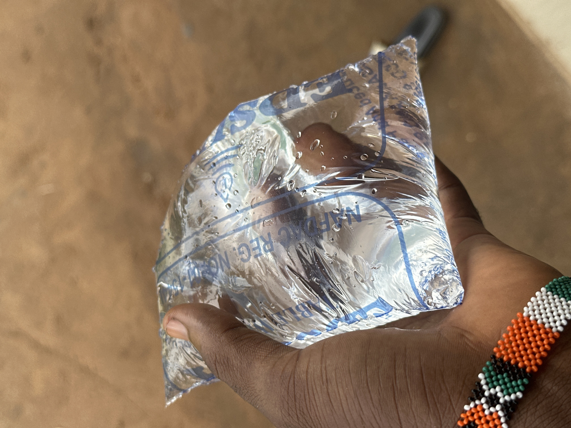 An arm and hand, adorned with a beaded bracelet featuring orange, white, green, and black beads, holds a transparent plastic bag filled with liquid. The bag is labeled with blue lettering; the phrase "NAFDAC REG. NO." is visible. Droplets of water cling to the bag's surface. The background is a blur of brown and tan, suggesting an outdoor or unrefined surface. The image likely represents a sachet of drinking water, a common sight in Nigeria. While the city of Ikot Ekpene is mentioned, there is no direct evidence in the image itself to confirm the location.