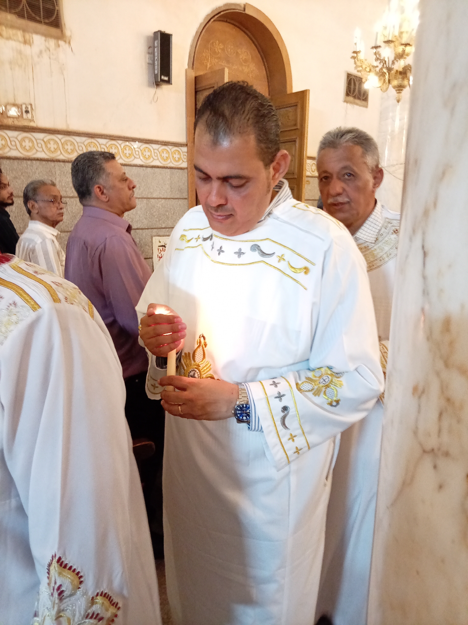 A medium shot captures a scene inside an ornate building, likely a church, with several people present. The central figure is a man, possibly a priest, wearing a white robe adorned with gold trim and intricate designs. He is holding a lit candle in his hands, his gaze directed downwards towards the flame. He is also wearing a blue watch.
Behind the priest are other men, their presence adding depth to the scene. One of them, positioned to the right of the priest, wears a similar white robe, suggesting he may also be a member of the clergy. In the background, other men are visible, some with glasses and wearing casual shirts.
The environment includes architectural elements, such as an archway, and a chandelier in the upper right of the frame. The walls are decorated with a patterned frieze, which extends behind the men. A pillar is visible on the right side of the frame, with beige or white surface.
The presence of religious attire and the lit candle strongly suggest a religious ceremony.
The location is Unknown, Unknown.
