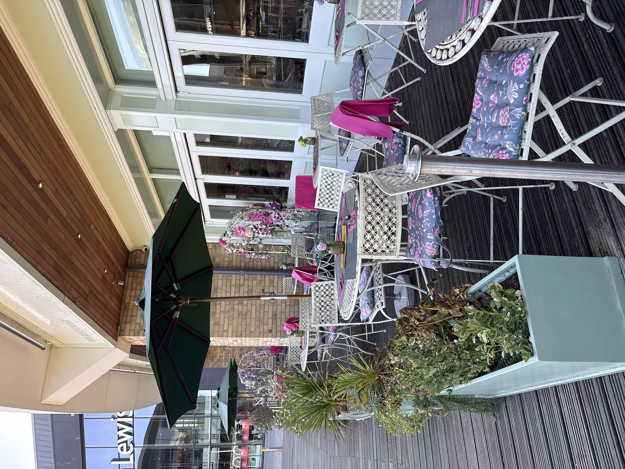 The image shows the outdoor seating area of a restaurant in Chelmsford, United Kingdom. The seating area is on a wooden deck with several white metal tables and chairs. The chairs have floral cushions in shades of pink and blue. Some tables have pink throws draped over them. There are green umbrellas providing shade. The restaurant building has large windows and doors, with a light green painted facade. Above the windows, a wooden overhang is visible. To the left of the seating area, a large glass-fronted building with "Lewis" signage is visible. The overall scene depicts a commercial establishment with an outdoor dining space.