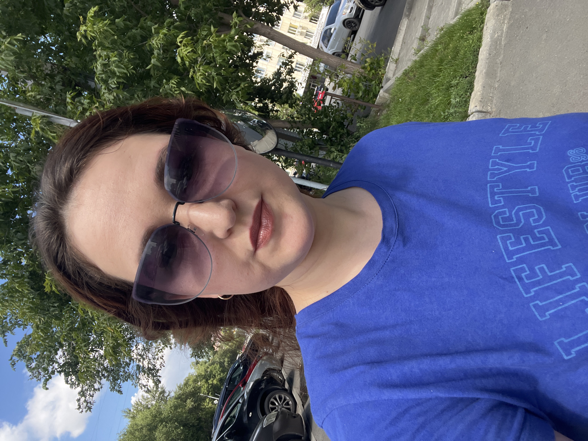 A woman in a blue t-shirt with sunglasses takes a selfie outdoors. She has dark brown hair and is wearing make-up. The background shows a street with parked cars, trees, and a building. The image is a street scene in Chelyabinsk, Russia.