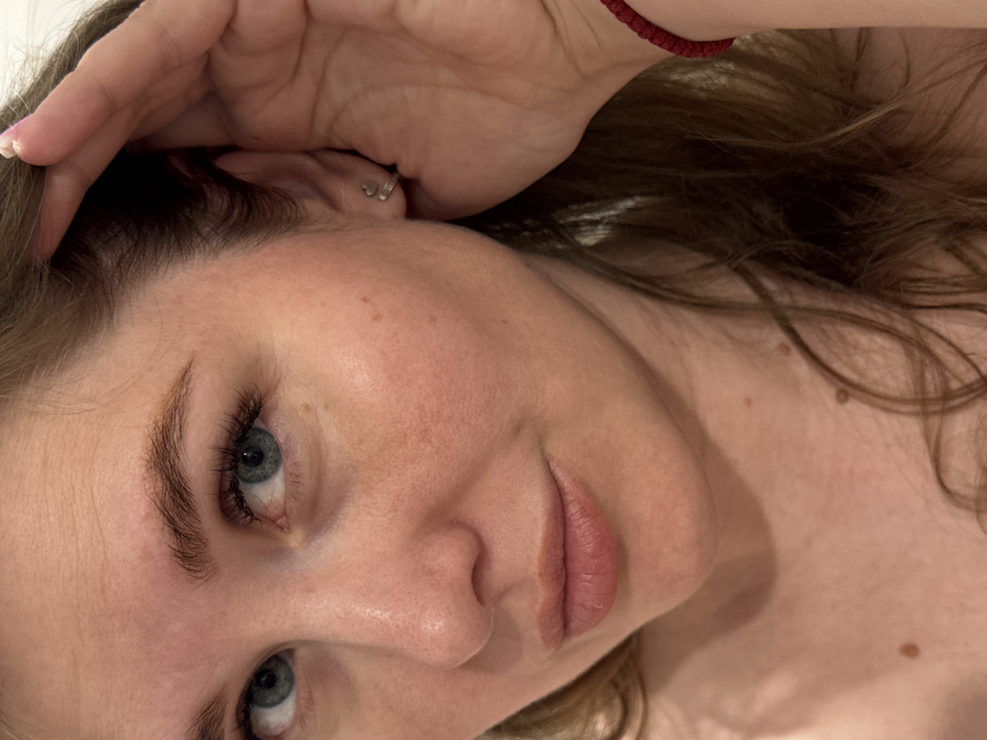 The image displays a close-up of a female subject, observed from the chest up, situated in Ферма, Russia. The subject has fair skin, blue-grey eyes with dark, full eyelashes, and medium brown hair that is parted and falls around her shoulders. Her lips are a natural pinkish-brown, slightly parted. Her nose exhibits visible pores, and a few small moles or freckles are present on her right cheek and upper chest. A silver-colored heart-shaped stud earring is worn in her right earlobe. Her right hand is raised to the side of her head, with fingers positioned near her temple and ear. A red string bracelet is visible on her left wrist. The background is indistinct and appears to be a plain, light-colored surface, indicating an indoor setting.