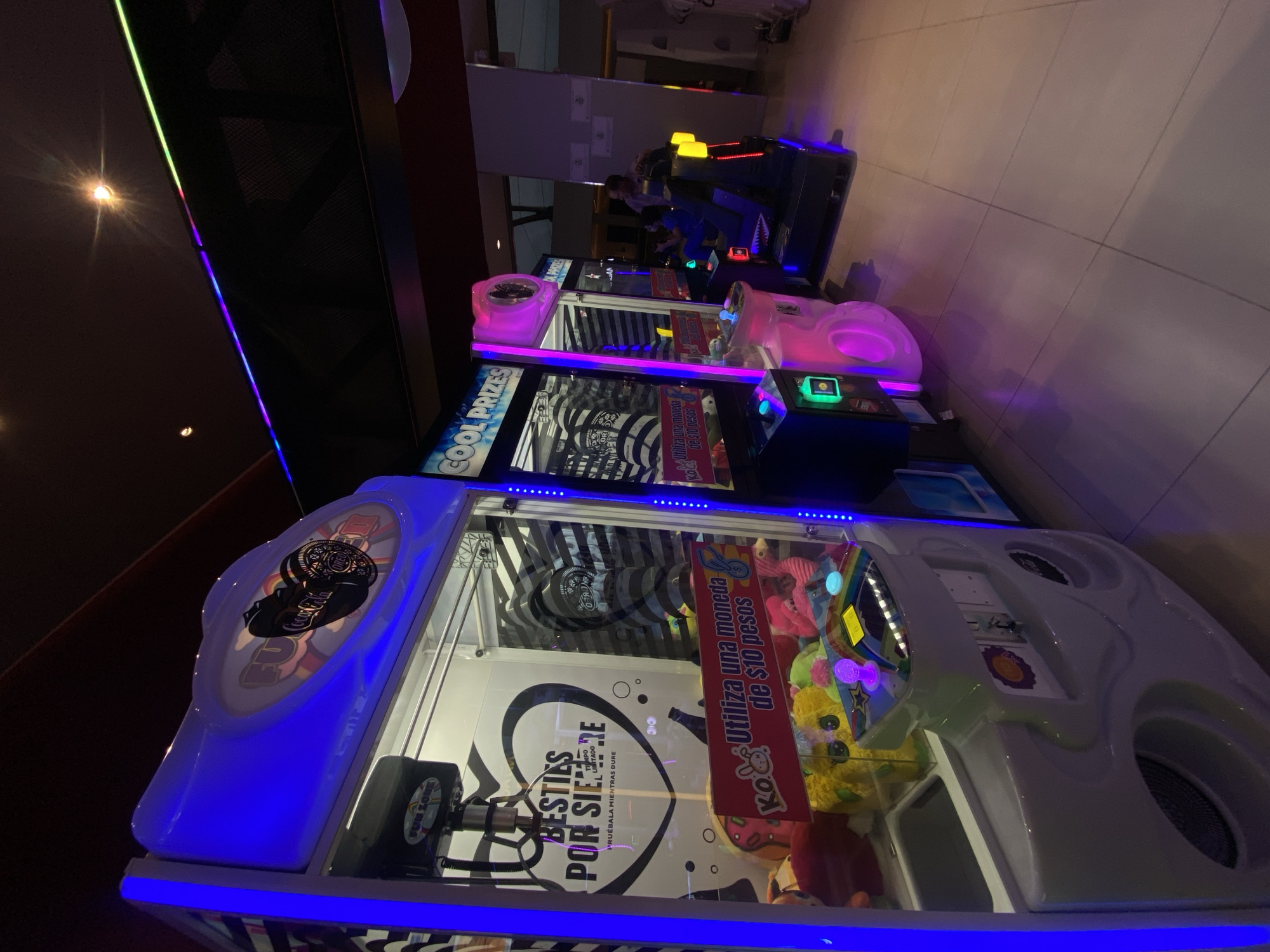 Here is a detailed description of the image:
The image depicts a brightly lit arcade featuring a row of claw crane machines. The nearest machine is a large, white-colored model with blue LED strip lighting. The interior of the machine is filled with plush toys, and a graphic on the front reads "Besties Por Siempre." Another machine further back has a pink top and is marked "Cool Prizes." In the background, there is a video game arcade with a person visible at one of the games. The location appears to be indoors, possibly in an entertainment venue.