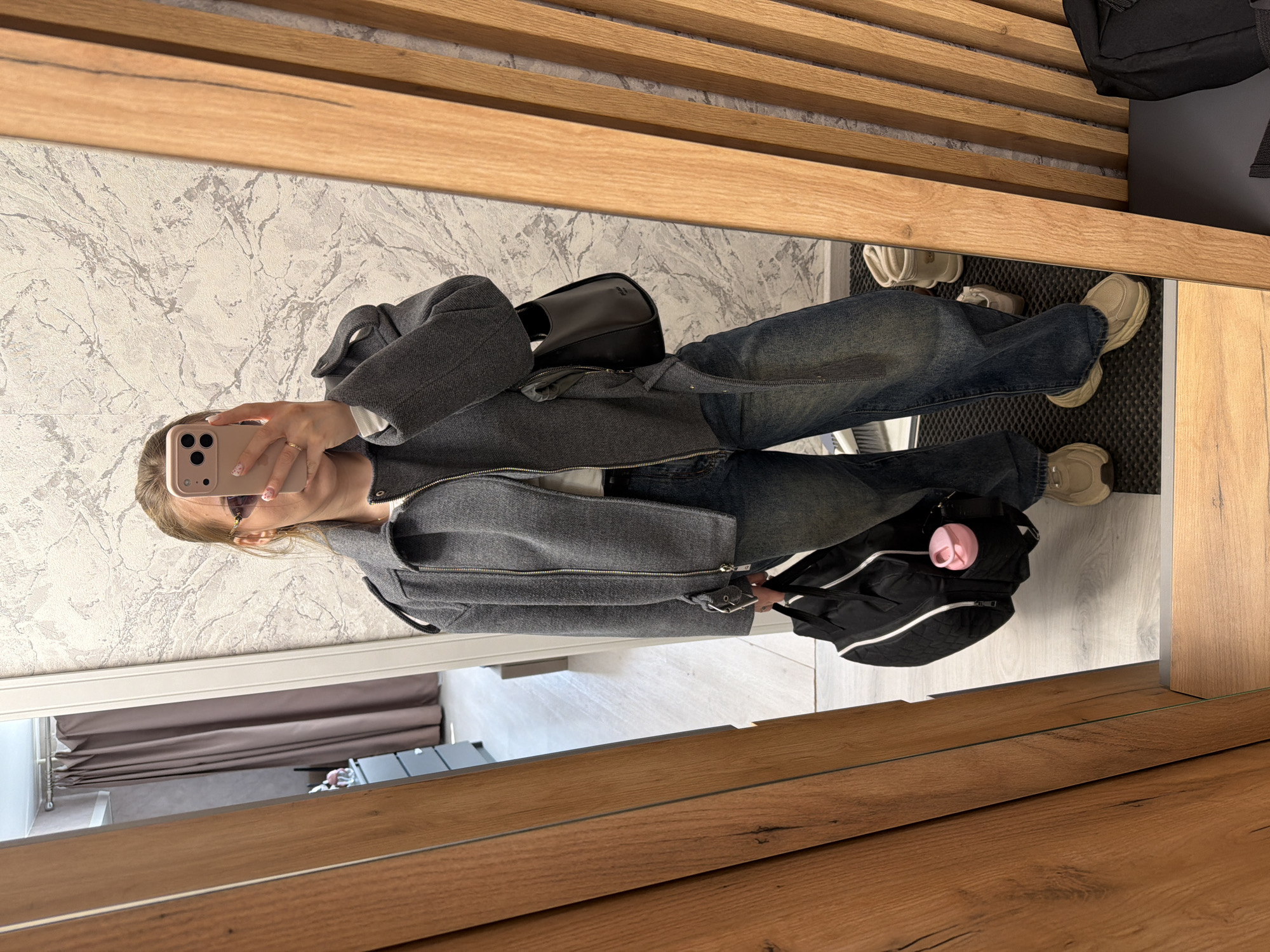 A female subject is depicted in a full-body mirror selfie, positioned within what appears to be a residential entryway or hallway in Reshetnikovo, Russia. She holds a light pink-cased smartphone with a triple camera array in her right hand, obscuring her upper face above the nose. She wears purple-tinted, rimless sunglasses visible just below the phone, a grey zippered jacket with shoulder epaulets and a buckle strap on the right sleeve, a white undershirt, dark blue relaxed-fit jeans, and beige chunky sneakers. A gold-toned ring is present on her right ring finger, and her fingernails display light-colored polish with small patterns. A small, black structured shoulder bag, possibly bearing a "PRADA" label, is carried on her right side. In her left hand, she holds a black duffel bag with white piping, featuring a pink water bottle in an exterior mesh pocket.
The immediate environment visible in the mirror includes a wall behind the subject covered in light-colored wallpaper with a grey and white marble-like pattern. The floor is light-colored wooden or laminate, with a black entrance mat directly in front of the subject. Vertical light brown wooden slats form part of the mirror's frame or an adjacent wall feature on the right. The reflection also shows an adjacent room through a doorway, revealing light-colored flooring, brown curtains hanging on a rod, a dark grey dresser with multiple drawers, and a small light-colored decorative object atop the dresser. A pair of light-colored boots and another pair of light shoes are present on the floor to the right of the subject's feet.