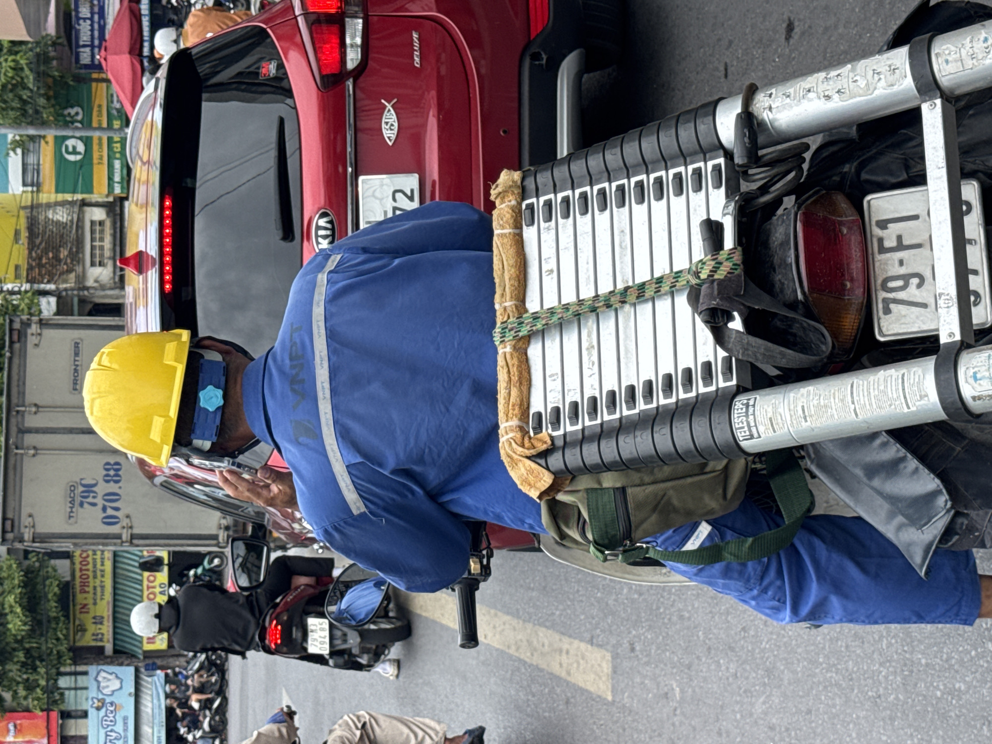 In Nha Trang, Vietnam, a person wearing a blue uniform and a yellow hard hat rides a motorcycle through moderate traffic. The motorcycle is carrying a folded ladder and a green bag. The rider is looking at their phone while navigating the road. In the background, a red Kia Sportage is visible, along with other vehicles and buildings. The scene appears to be a typical day on a busy Vietnamese street.