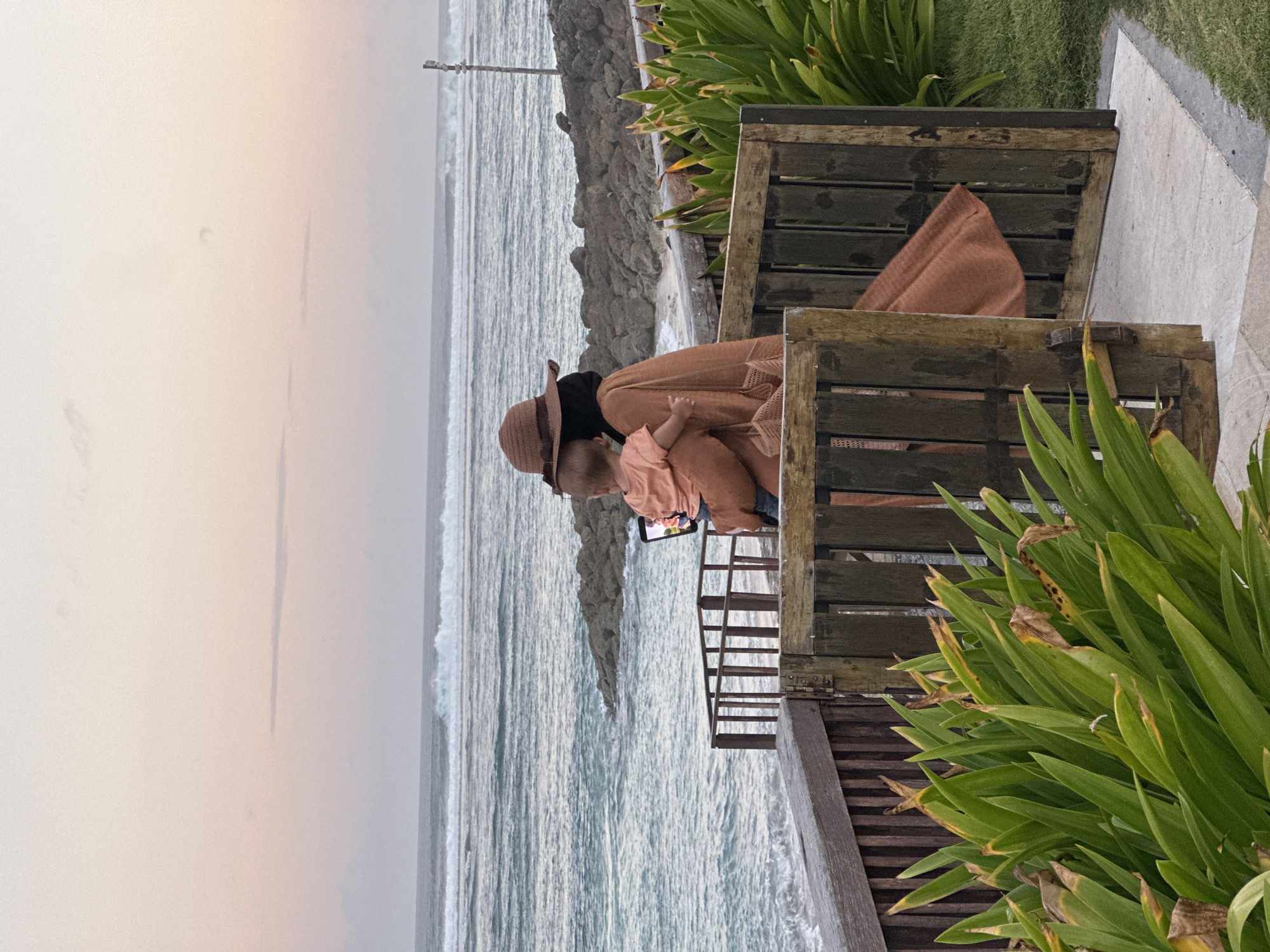 In Kuta Selatan, Indonesia, a person wearing a wide-brimmed hat and an earth-toned outfit is holding a baby. The person is facing away from the camera, looking out at the ocean where waves are breaking. They are holding a smartphone, seemingly capturing a photo or video of the scene. In the foreground, there are lush green plants and a wooden fence with a gate. The sky is a soft gradient of pink and orange, indicating either sunrise or sunset. The overall atmosphere is peaceful and serene, with the natural beauty of the beach and ocean as the backdrop.