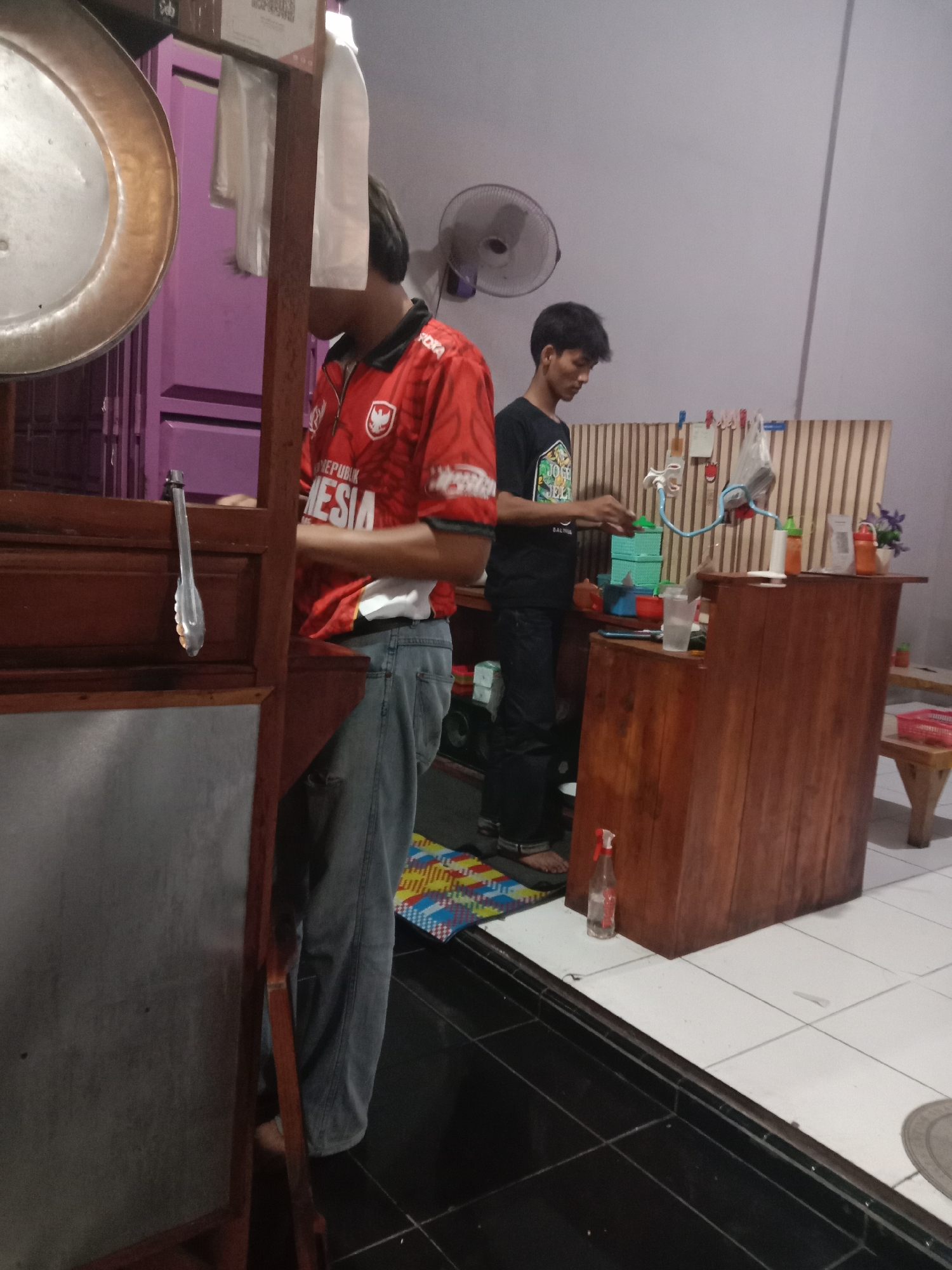 The image depicts a scene inside a food stall or small eatery in Cangkring, Indonesia. Two young men are present, appearing to be employees. The man on the left, wearing a red and white Indonesian football jersey and blue jeans, stands behind a wooden counter. He is facing away from the camera, and his head is turned slightly to his right. A pair of metal tongs hangs from the side of the counter.
The man on the right, wearing a black t-shirt and dark pants, stands behind another wooden counter. He is facing left and appears to be engaged in preparing food or drinks, with his hands positioned over a collection of green plastic containers. A glass of liquid, a plastic bottle with a spray nozzle, and several other items are visible on the counter. A white-board with various clips and papers is mounted on a striped partition behind him.
The environment appears to be an indoor, dimly lit space with tiled flooring. A large metal disc, possibly a part of cooking equipment, is visible on the far left. In the background, a purple cabinet or wall is visible. The overall atmosphere suggests a working commercial environment.