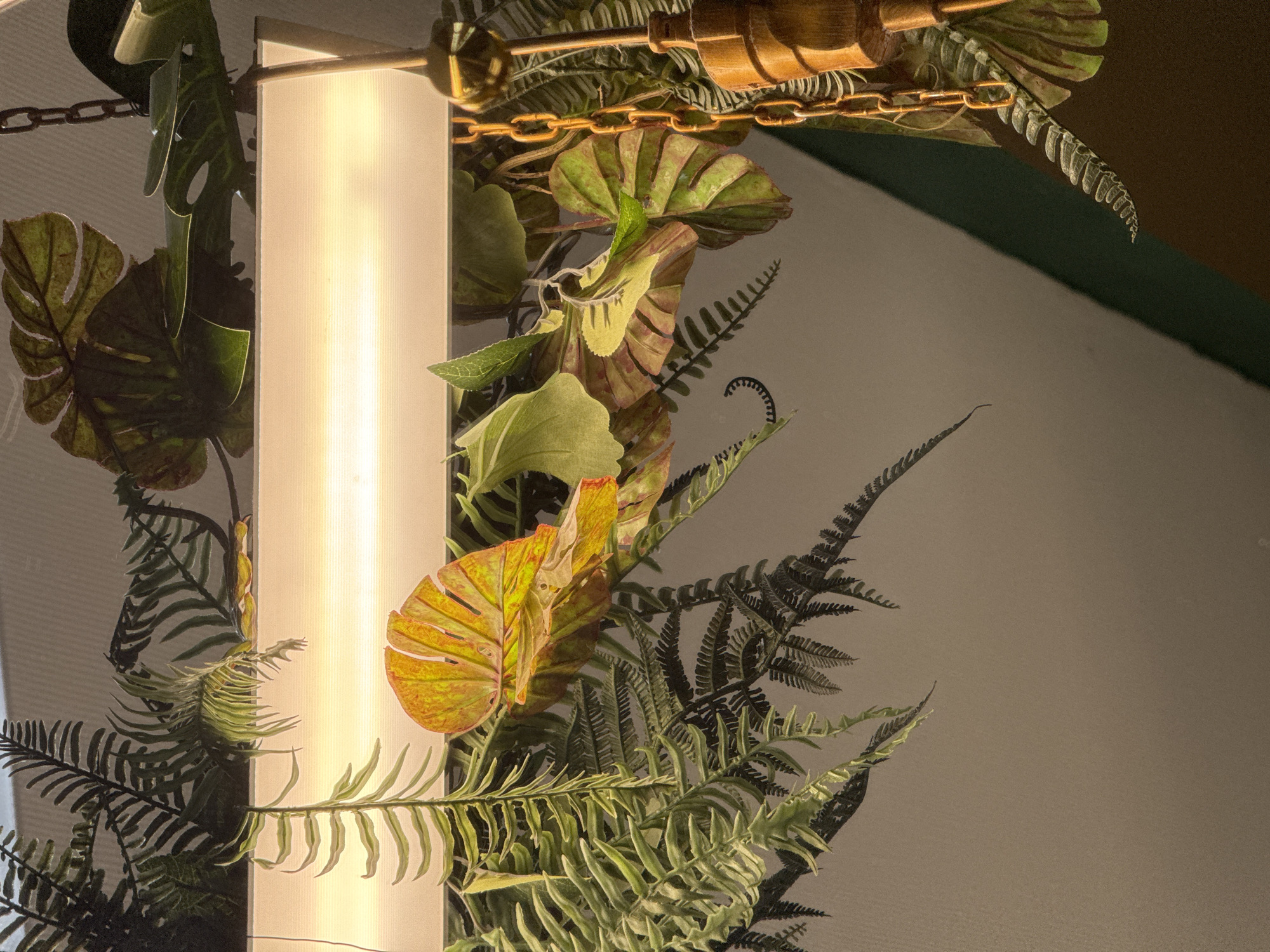 The image depicts an indoor scene focused on a horizontal, rectangular lighting fixture, likely fluorescent or LED, emitting a bright, diffuse white-yellow light. The fixture is surrounded and partially obscured by a dense arrangement of artificial foliage. This foliage includes numerous green fern fronds with pinnate leaves and larger, broad tropical-style leaves, some of which exhibit green coloration while others feature prominent yellow and reddish-orange hues, suggesting faux aging or specific plant types. On the upper right, a segment of a golden-colored metal chain is visible, descending vertically. Below this chain, a decorative element, possibly made of wood or brass, is partially observed. The background consists of a light-colored, uniform surface (likely a ceiling or upper wall) and, in the lower right quadrant, a darker, diagonally angled surface, possibly a feature wall or architectural detail. No individuals are present. The specific location within the city of Chaikovsky, Russia, cannot be determined from the provided visual information.