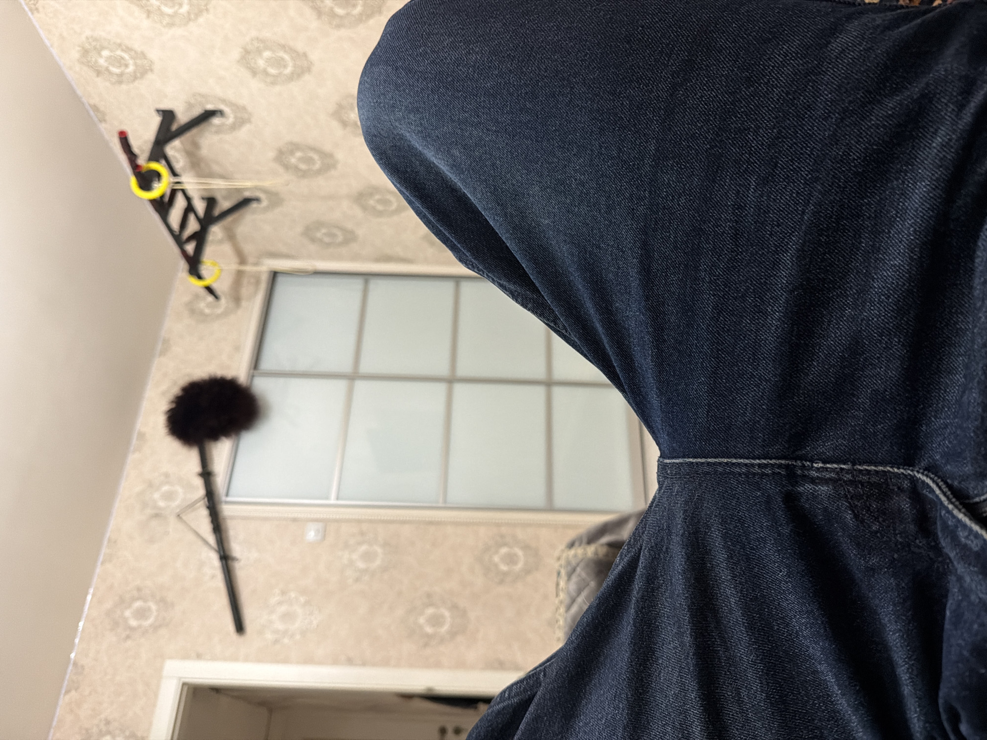 The image shows a first-person perspective of a person sitting down, with their dark blue denim-clad legs prominently in the foreground. The background consists of a wall with a patterned wallpaper. Mounted on the wall are two objects: a long, thin bar with a dark, fluffy object attached to one end, possibly a duster or microphone boom, and a black pull-up bar with yellow rings and hanging straps. To the left, a doorway leading into another room is partially visible. In the center of the background, a large, rectangular sliding door with a grid pattern is present. The lighting suggests an indoor environment. The location is an unknown city in Russia.