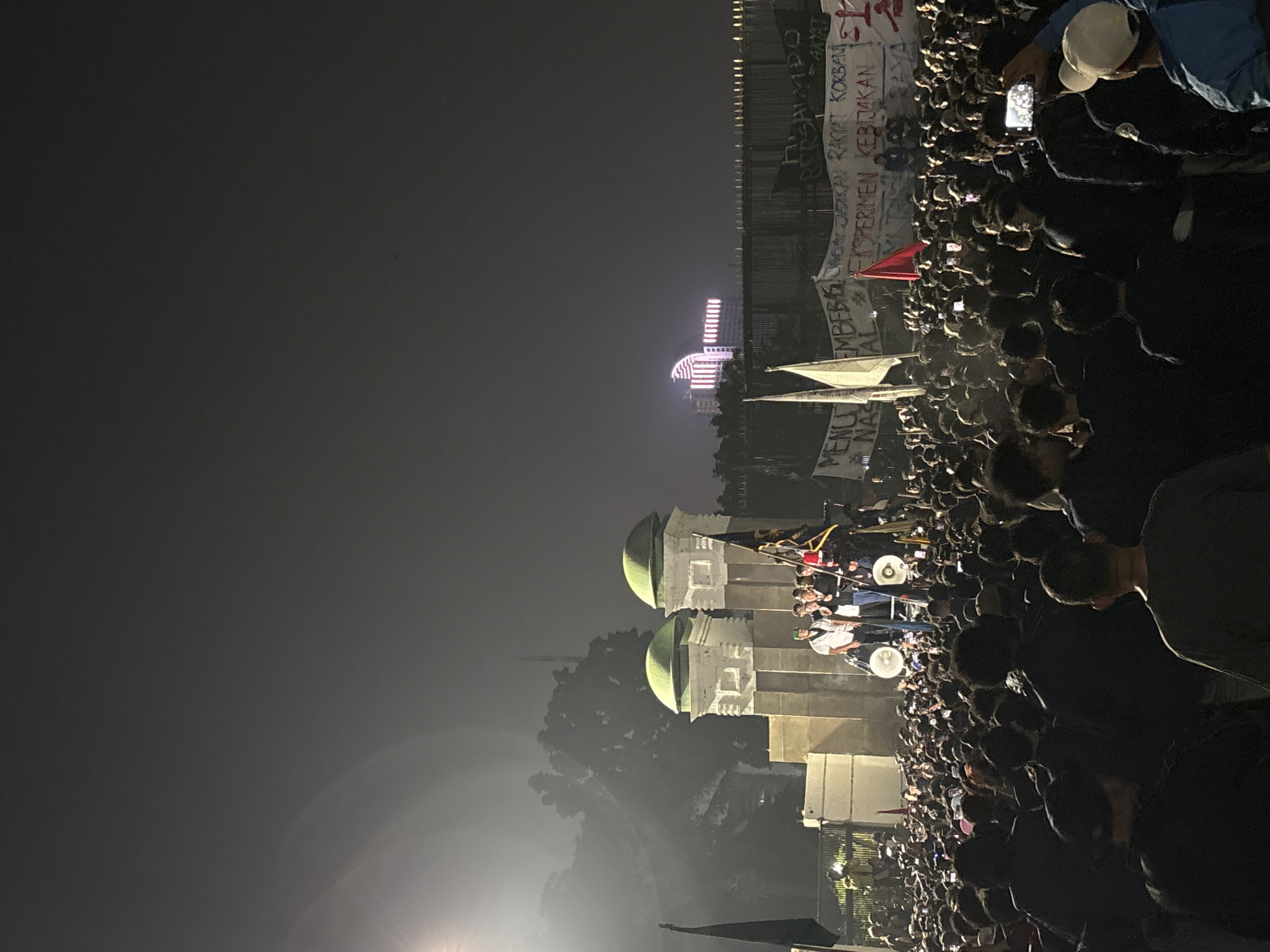 The image depicts a nighttime gathering of a large crowd of people in Central Jakarta, Indonesia. The scene appears to be outdoors, with a dark, hazy sky suggesting nighttime conditions and possibly fog or light pollution. In the background, a modern building with illuminated windows is visible, hinting at an urban setting. Dominating the foreground and mid-ground are the assembled people, many holding up their mobile phones, suggesting they are documenting or engaged with an event.
A prominent feature in the mid-ground is a structure with two rounded green domes atop light-colored buildings, possibly a historical or significant landmark. Beside this structure, a group of people are gathered around what appears to be a platform or trolley, with some individuals standing and others seated. Flags are visible within the crowd and near the platform.
Several banners with text are present. One banner, partially visible, reads "MEMBERI KORBAN" and "KEBIJAKAN", suggesting themes of sacrifice and policy. Another banner displays text in what appears to be Indonesian. The overall atmosphere appears to be one of collective engagement, possibly a protest, rally, or public event, given the dense crowd and the banners. The lighting is dim, with the illumination coming from the building in the distance and the mobile phone screens. The time of day is clearly night.