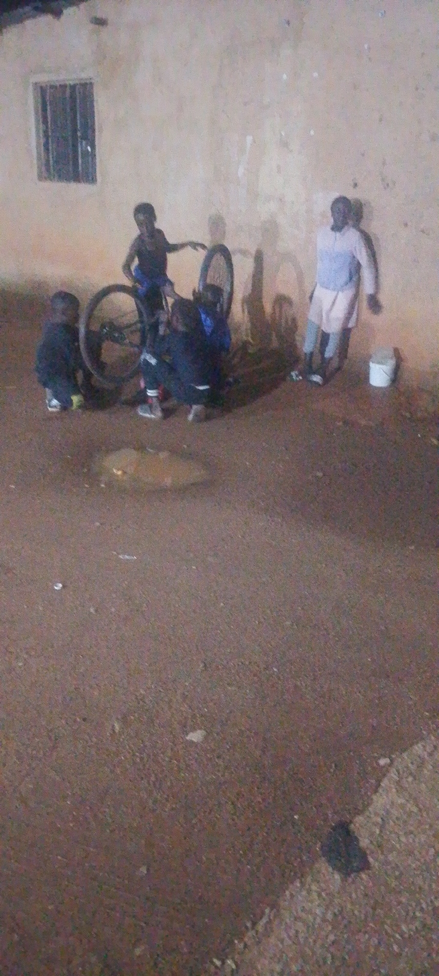 In Gura Top, Nigeria, a group of individuals are gathered around a bicycle against a textured, light-colored wall. A small window with bars is visible on the left side of the wall. The ground is a reddish-brown dirt. In the center, a bicycle is propped up, and two boys are crouched down, appearing to be working on it. One boy is wearing a dark shirt and blue shorts, while the other wears a dark jacket with white accents on his pants. To the left of the bicycle, a third child is standing and observing. Further to the right, a man in a light blue shirt and white shorts stands near a white bucket. Shadows are cast on the wall from the figures and the bicycle, suggesting the lighting source is to the left of the scene. The overall impression is of an informal gathering focused on the bicycle, possibly during maintenance or repair.