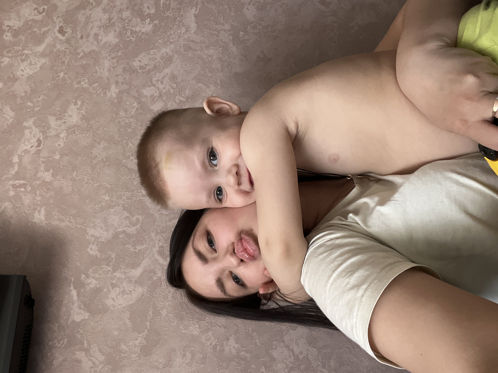 An indoor portrait, originating from the city of Марковский, Russia, depicts an adult female and a young male child. The background consists of a light-brown wall with a mottled, textured pattern. In the upper left quadrant, a dark rectangular wall-mounted device with visible grilles is partially present.
The adult female, positioned on the left, has long dark hair, light eyes, and fair skin. She wears a light-colored short-sleeved t-shirt and a necklace with a small pendant. Her left hand is raised to her cheek/jawline. She is looking directly at the camera with puckered lips, forming a "duck face". A ring is visible on her left ring finger.
The young male child, positioned to the right of the adult and embracing her, has short reddish-blonde hair, light eyes, and fair skin. He is shirtless. A yellowish bruise is present on his forehead above his right eye. He holds the adult with his right arm around her neck/shoulder and his left arm across her front. He displays a slight smile and looks slightly towards the viewer's right. A small yellow object with a black component is partially visible in the bottom right corner, beneath the adult's right hand.