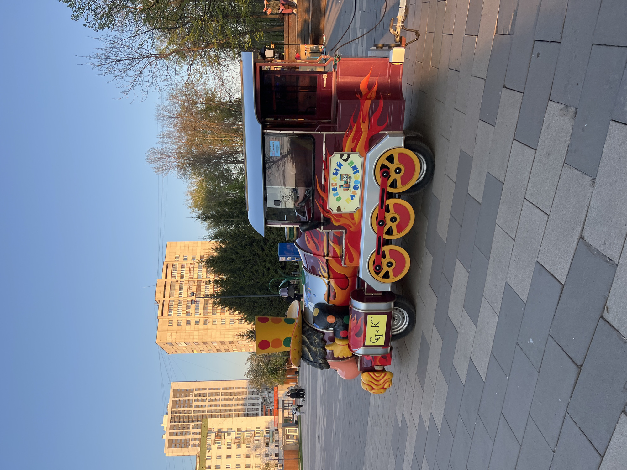 The image depicts a colorful children's train parked on a paved walkway in Ufa, Russia. The train is designed with cartoonish features, including faux steam-engine details and a clown-like figure at the front. The train has decorative flame patterns along the sides. In the background, there are apartment buildings and trees, typical of an urban environment. The sky is clear and blue, suggesting a sunny day. There are a few people walking in the distance. The walkway is constructed from gray paving stones. The orientation of the image is vertical.