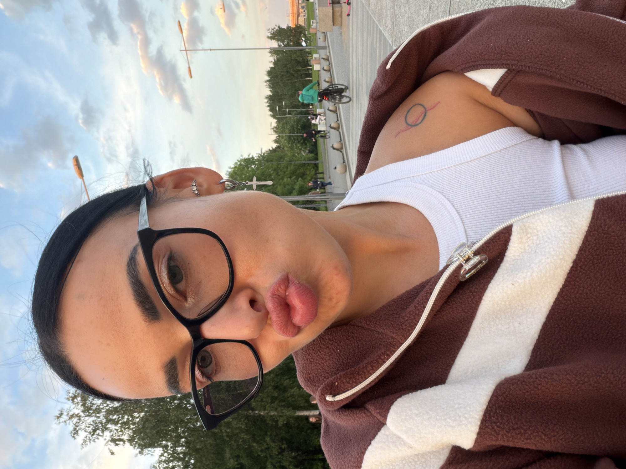 A selfie image, rotated 90 degrees clockwise, features a person captured outdoors in what appears to be late afternoon or early evening light, suggested by the warm hues in the sky. The subject, positioned with their head on the left and body extending to the right, has dark hair pulled back and wears large black-framed glasses. They are making a "duck face" or "kissy face" with pursed lips, giving a playful or confident expression.
The person is wearing a white ribbed tank top layered under a brown fleece jacket with wide white stripes. Visible accessories include multiple earrings: a silver cross dangling from the right ear, a hoop, and studded earrings. A small, stylized tattoo, featuring a red line underneath a blue-green circle, is visible on their upper left arm.
The background depicts an outdoor public space, likely a promenade or park in Saint Petersburg, Russia. Trees, streetlights, and distant figures of other people are visible, including one person riding a bicycle. The sky is partly cloudy with soft, scattered clouds, complementing the gentle lighting. No visible text is present in the image.