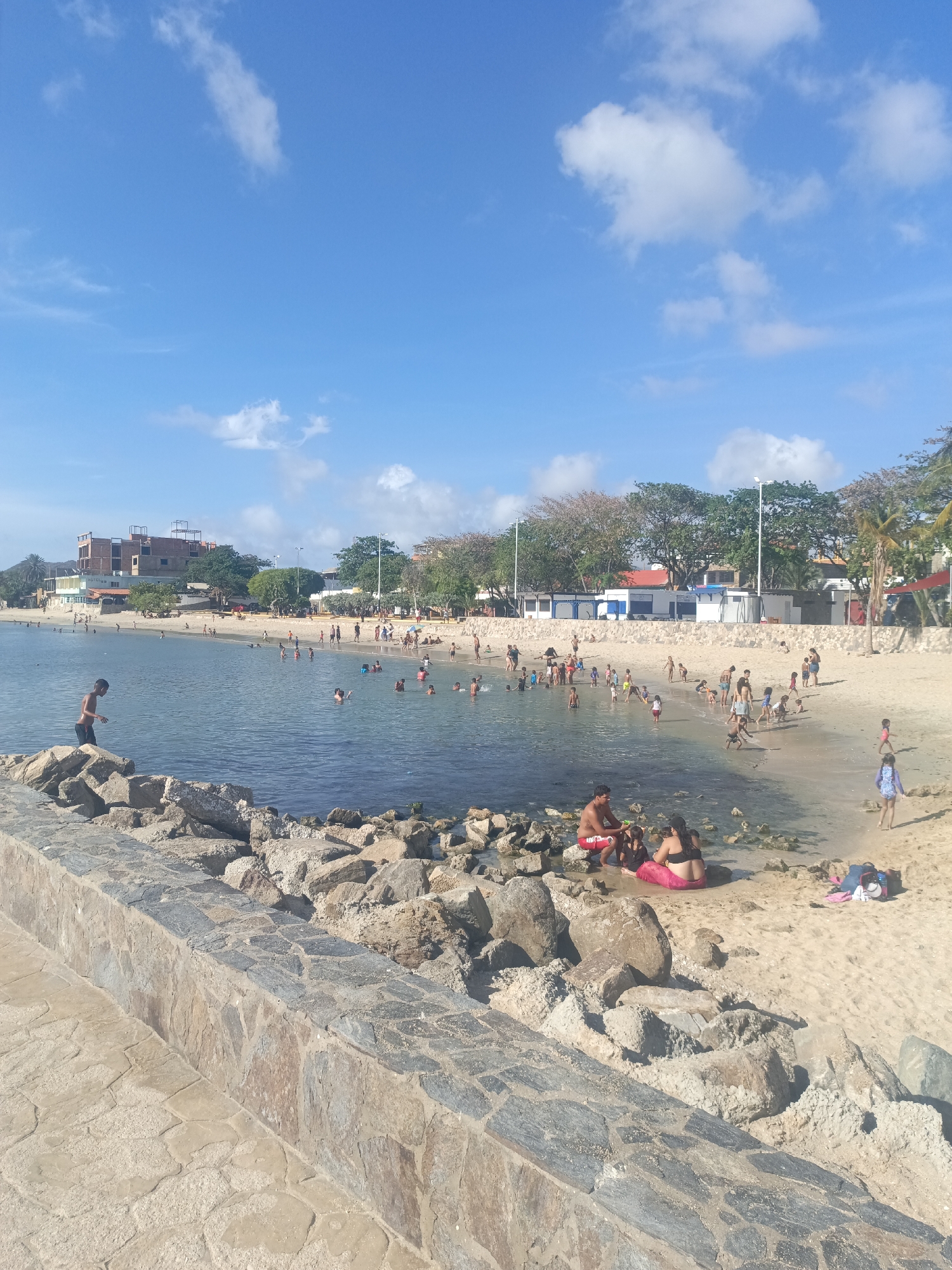This is a daytime shot of a beach in Juan Griego, Venezuela. A seawall, composed of stone and cement, dominates the foreground. Beyond the seawall, the beach is populated with numerous people, adults and children. Some are in the water, swimming or wading, while others are on the sand, playing or relaxing. A family group, consisting of adults and children, are sitting on the beach near the seawall. Buildings are visible in the background, along with trees and street lamps. The sky is blue with scattered clouds.