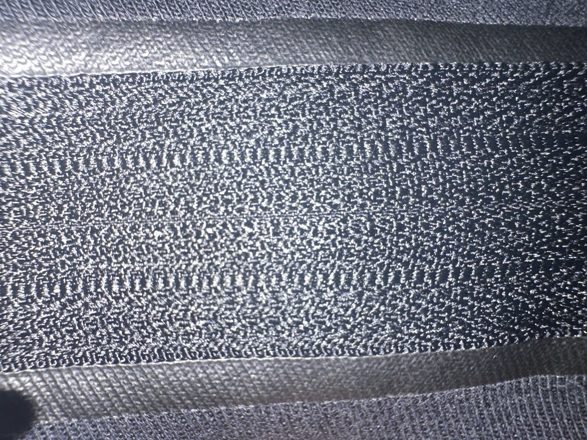 This is a close-up, abstract image of a dark, textured material, possibly fabric or a woven surface. The dominant colors are dark gray and black, with bright white highlights scattered across the surface, suggesting a rough or slightly reflective texture. The material appears to be layered, with distinct horizontal bands. One band, in the center, has a more intricate, granular texture with a shimmering quality. Above and below this central band are smoother, darker bands. The lighting creates strong contrasts, with bright reflections on the textured areas and deep shadows elsewhere. There is no discernible scene, setting, or identifiable objects. No people are visible, and no activity or event is taking place. There is no visible text in the image. The location context of Letterkenny, Ireland, is not visually represented in the image itself.