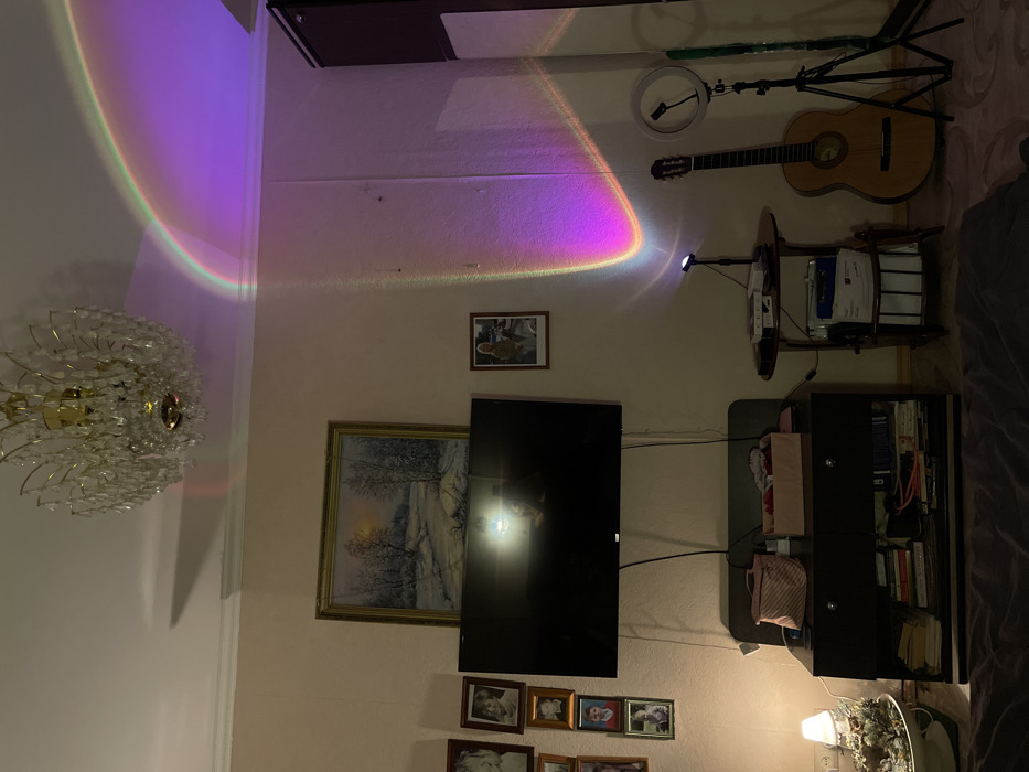 A living room features a television mounted on the wall, flanked by an ornate painting on the left and a bookshelf to the right. Below, a collection of framed photos decorates the wall. Above, a guitar rests near a ring light on a tripod. A curved, colorful light effect shines across the upper wall, emanating from a projector. A crystal chandelier hangs from the ceiling. A small two-tiered table also is present.