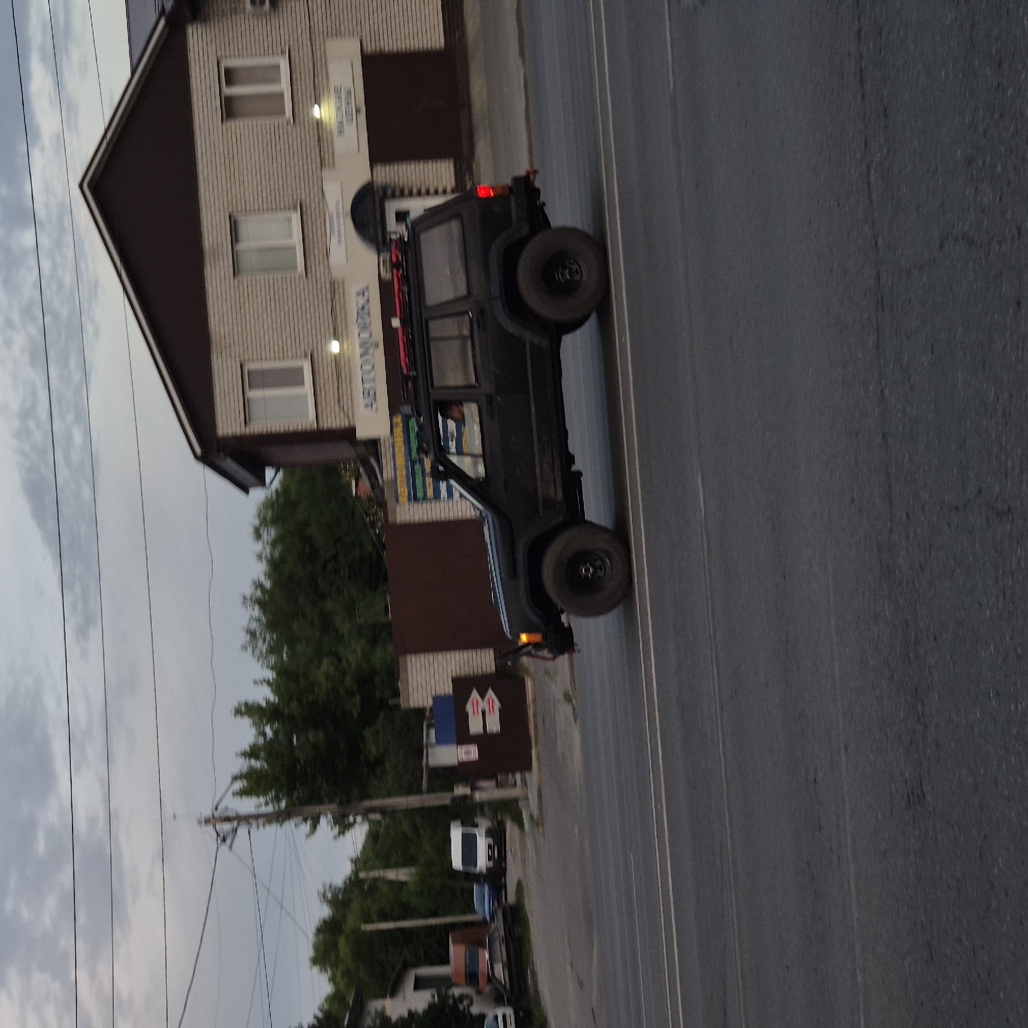A dark-colored Jeep Cherokee is captured in motion, driving on a paved road. The vehicle is positioned in the right half of the frame, angled slightly towards the viewer's left. The Jeep has a roof rack with red accents. Its front left tire is prominently visible, and its brake light is illuminated red on the right rear. The road appears to have multiple lanes, indicated by white lane markings. In the background, a multi-story building with light-colored brick siding and a dark-colored pitched roof is visible. Above the building, power lines crisscross the cloudy sky. To the left of the building, a row of trees forms a green backdrop. Further left, another building, possibly a smaller house or structure, is partially visible. The scene suggests an urban or suburban environment.